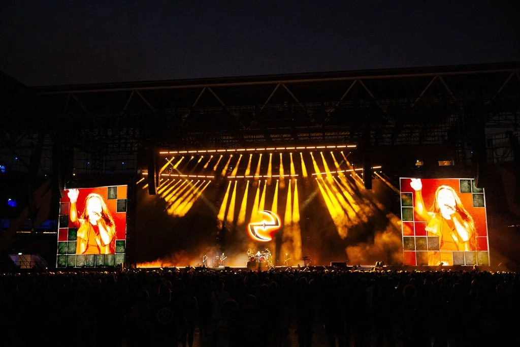 A large concert stage with bright yellow lights and screens showing a singer, set at night with a crowd in the foreground.