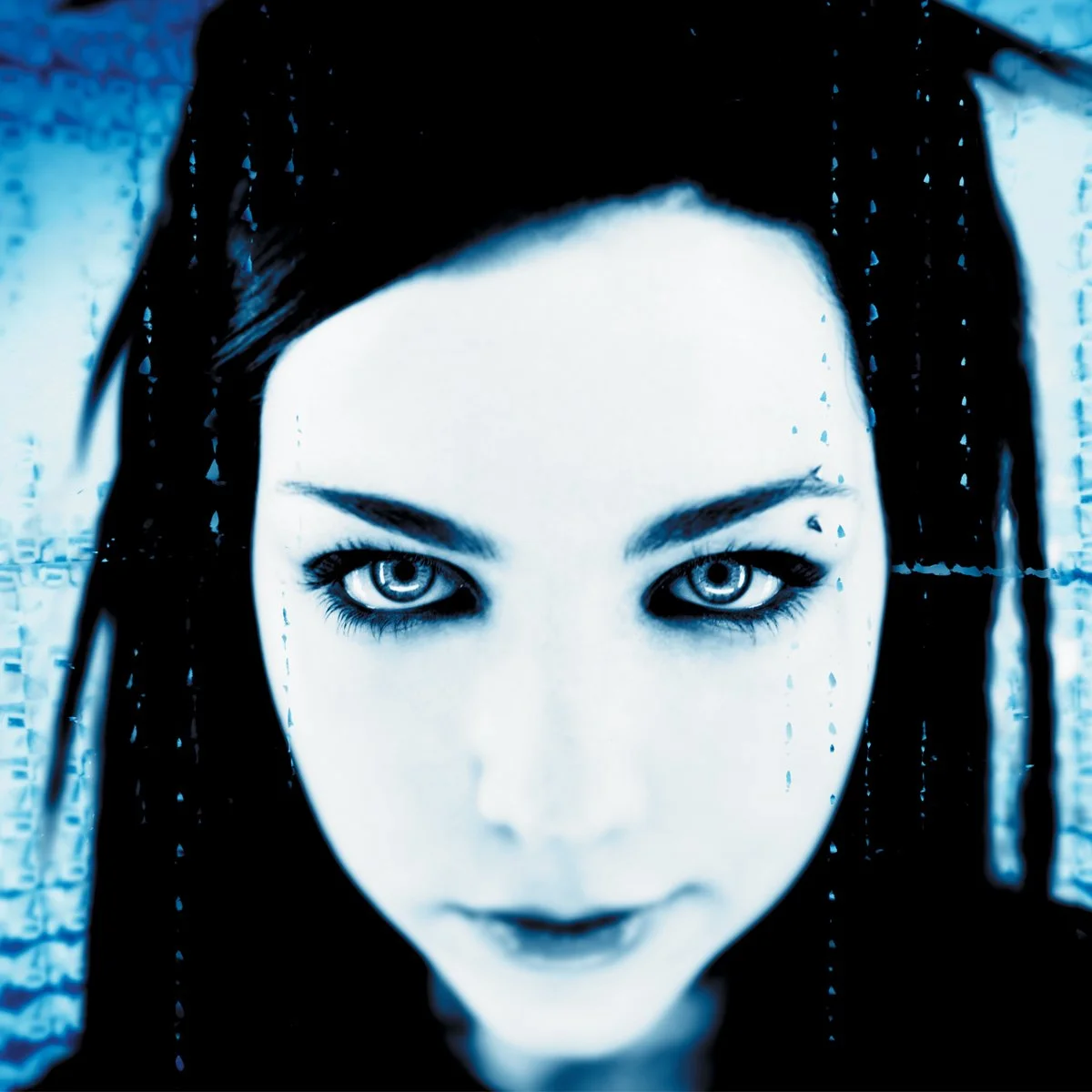 Close-up of a woman's face with intense blue eyes, dark hair with braided streaks, and gothic makeup, looking directly at the camera.