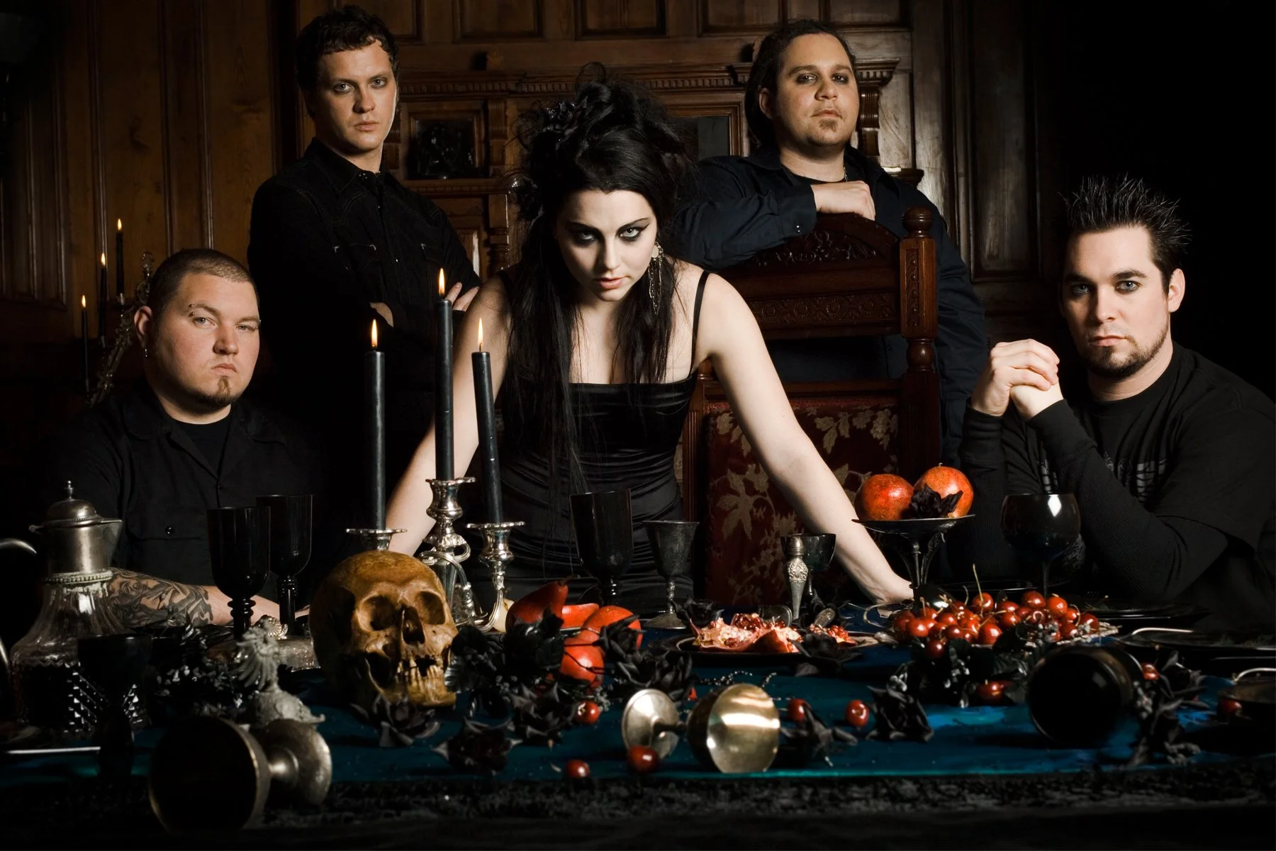 A gothic-themed group portrait of five people seated at a dark ornate table with candles, skull, and food items, in a wood-paneled room.