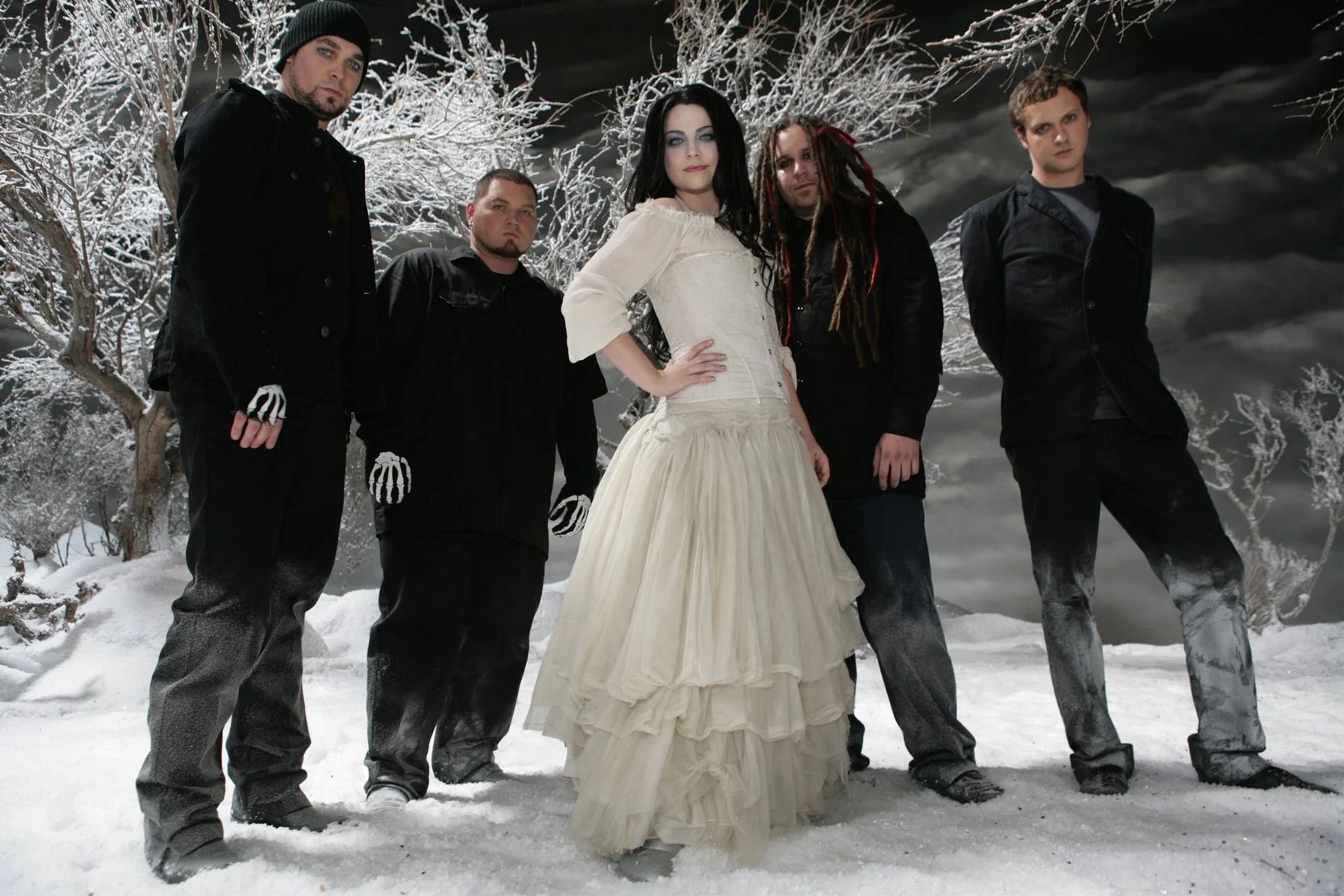 A woman in a long, cream-colored dress and five men in dark clothing standing in a snowy outdoor setting with leafless trees and a dark sky in the background.