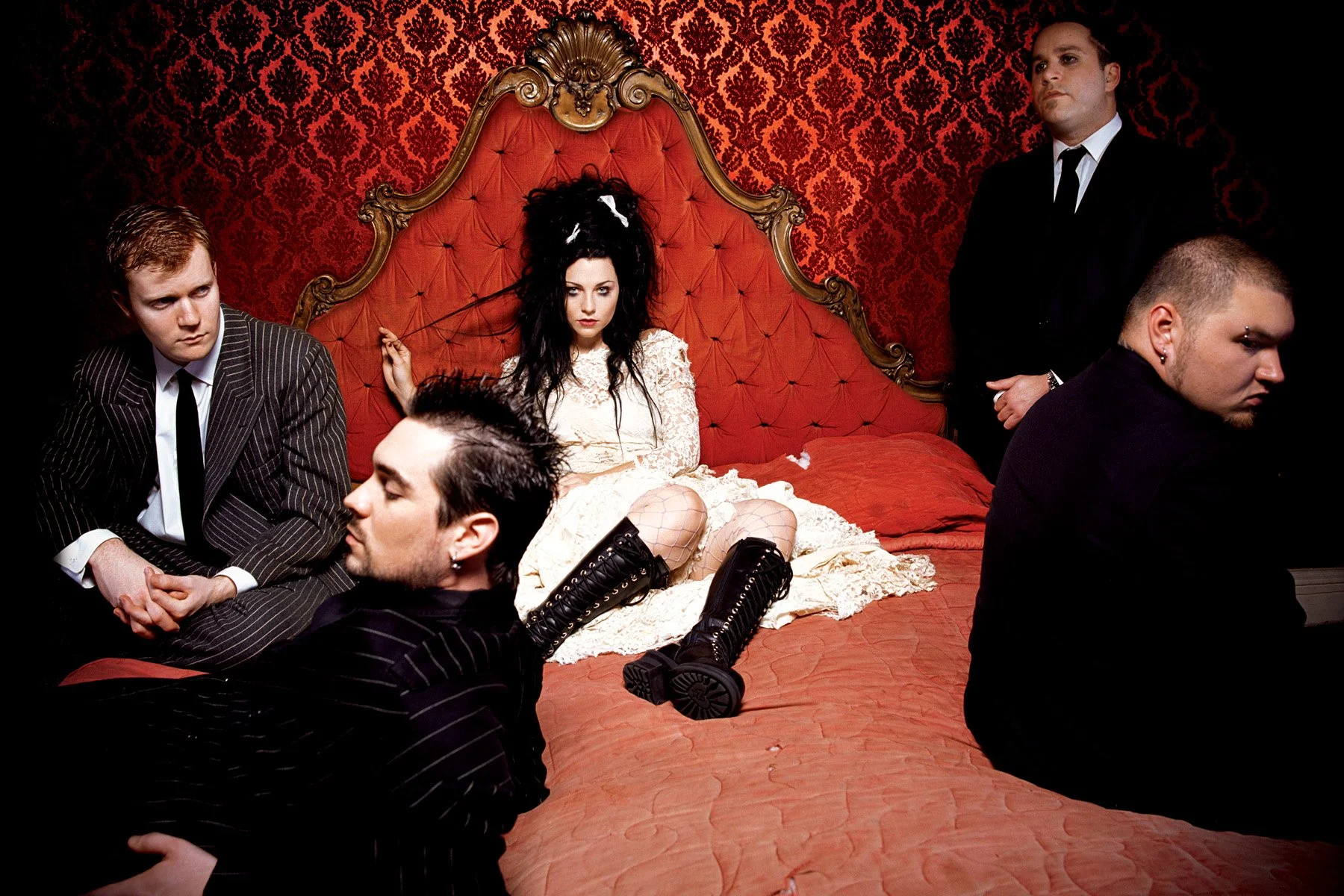 A woman with black hair and gothic makeup sitting on a red velvet bed with ornate gold trim, surrounded by four men in dark and striped clothing in a room with red and black damask wallpaper.
