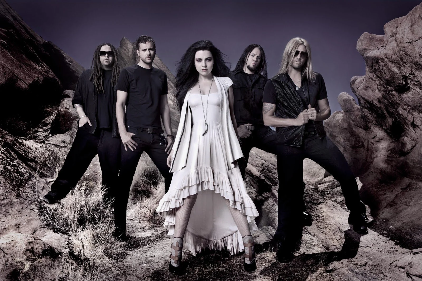 A band of five members posing in a rocky desert landscape, with mountains in the background; the woman in the center wears a white dress, while the men around her wear black clothing, some with sunglasses and tattoos.