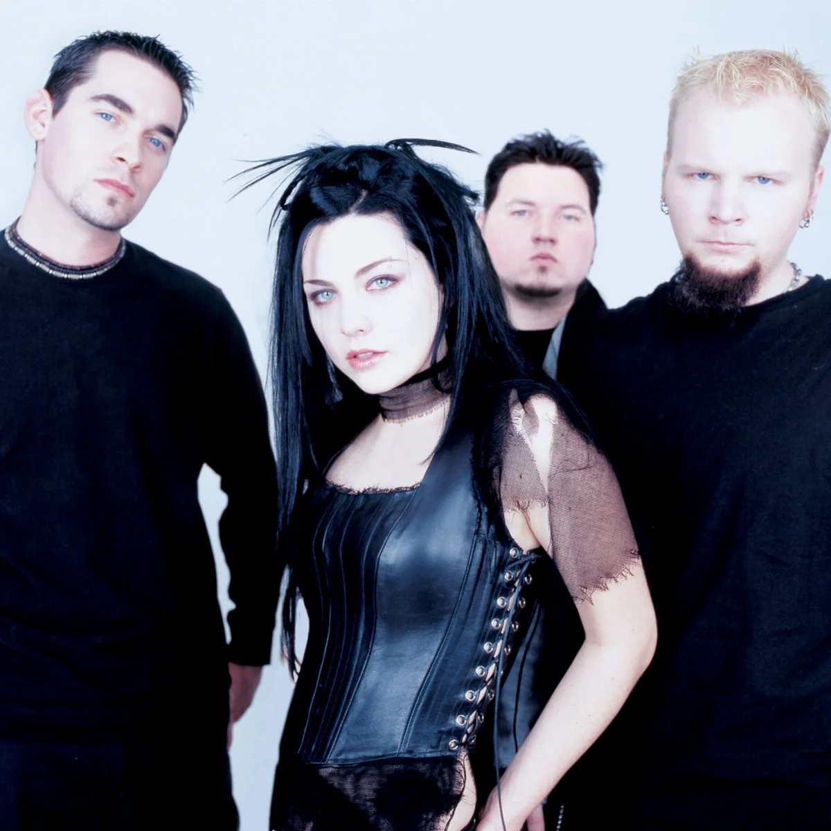 Four band members posing against a light background, with the woman in the center wearing a black leather corset and distressed mesh top, and the men dressed in black with varying hairstyles and facial expressions.
