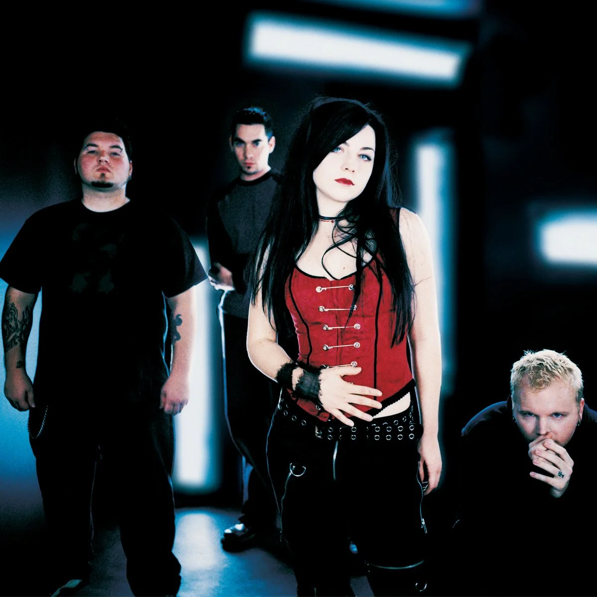 A group of four people posing in a dark room with blue lighting, with a woman in the center wearing a red corset and black pants, and three men behind or beside her.