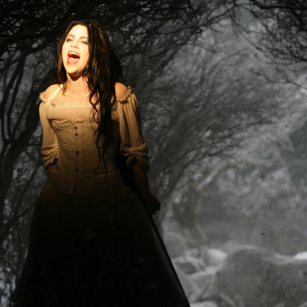 A woman with long dark hair singing or shouting outdoors at night, with bare tree branches and snow in the background.