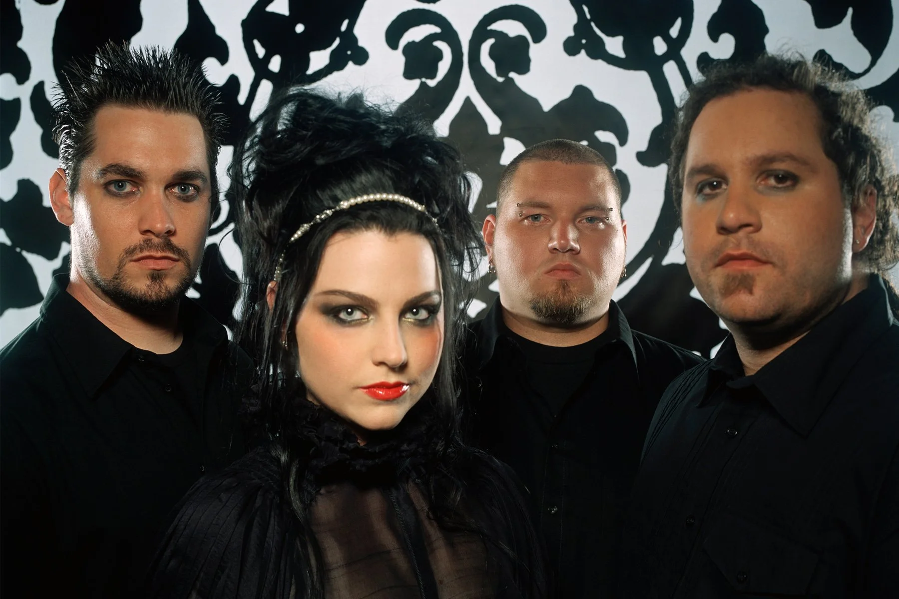 Group portrait of four band members with dark clothing and makeup, standing in front of a black and white abstract background.