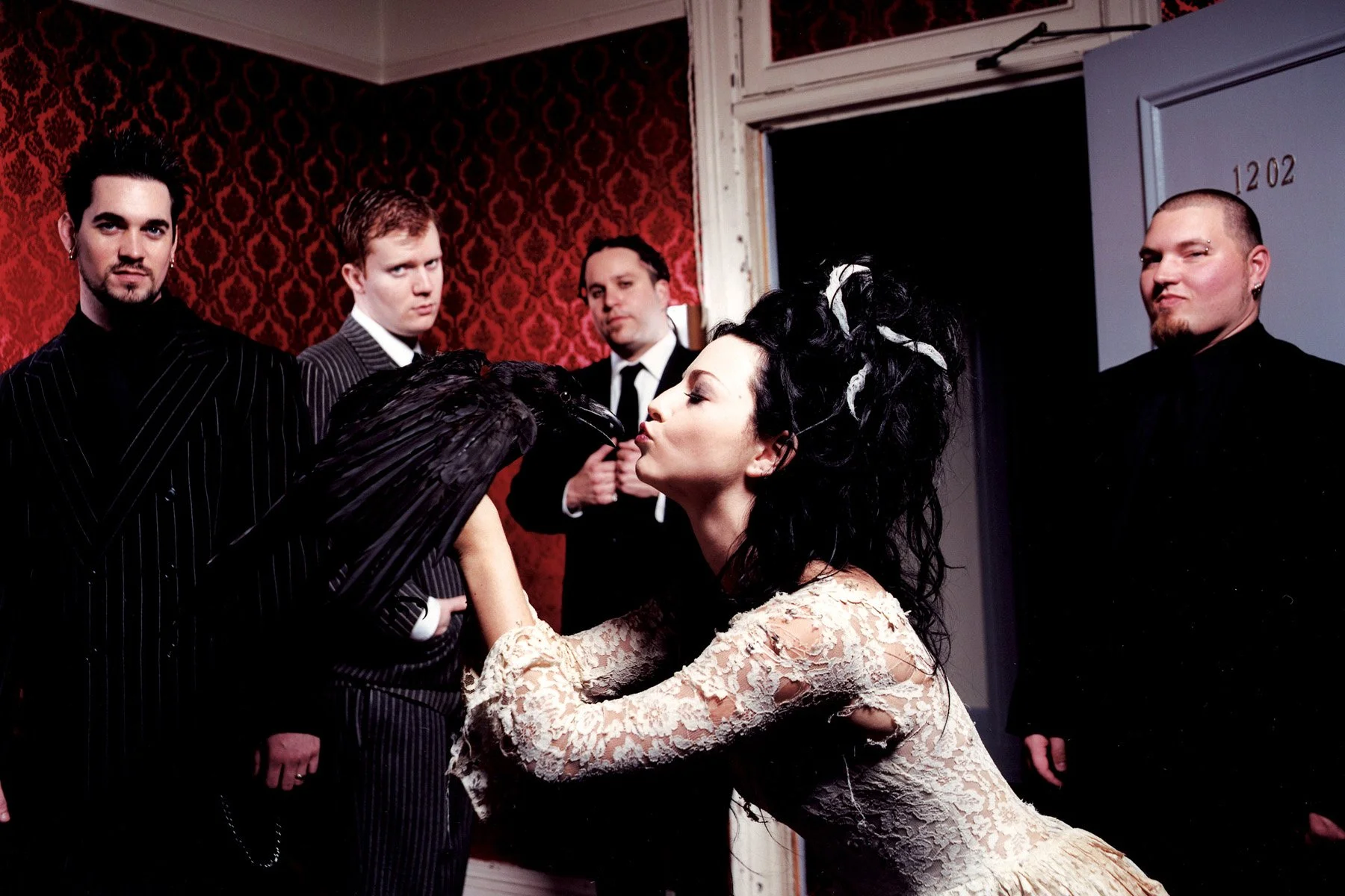 A woman with dark, curly hair and pale skin is kissing a black crow. Four men in black suits are standing nearby in a room with red patterned wallpaper, watching the scene.