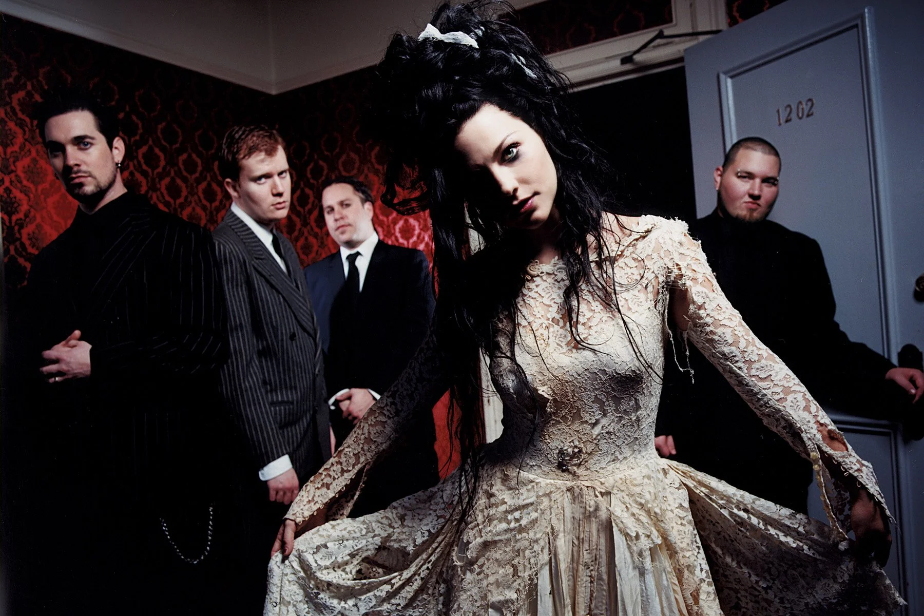 A woman with long black hair wearing a vintage lace dress in front of four men in suits in a dimly lit room with red wallpaper.