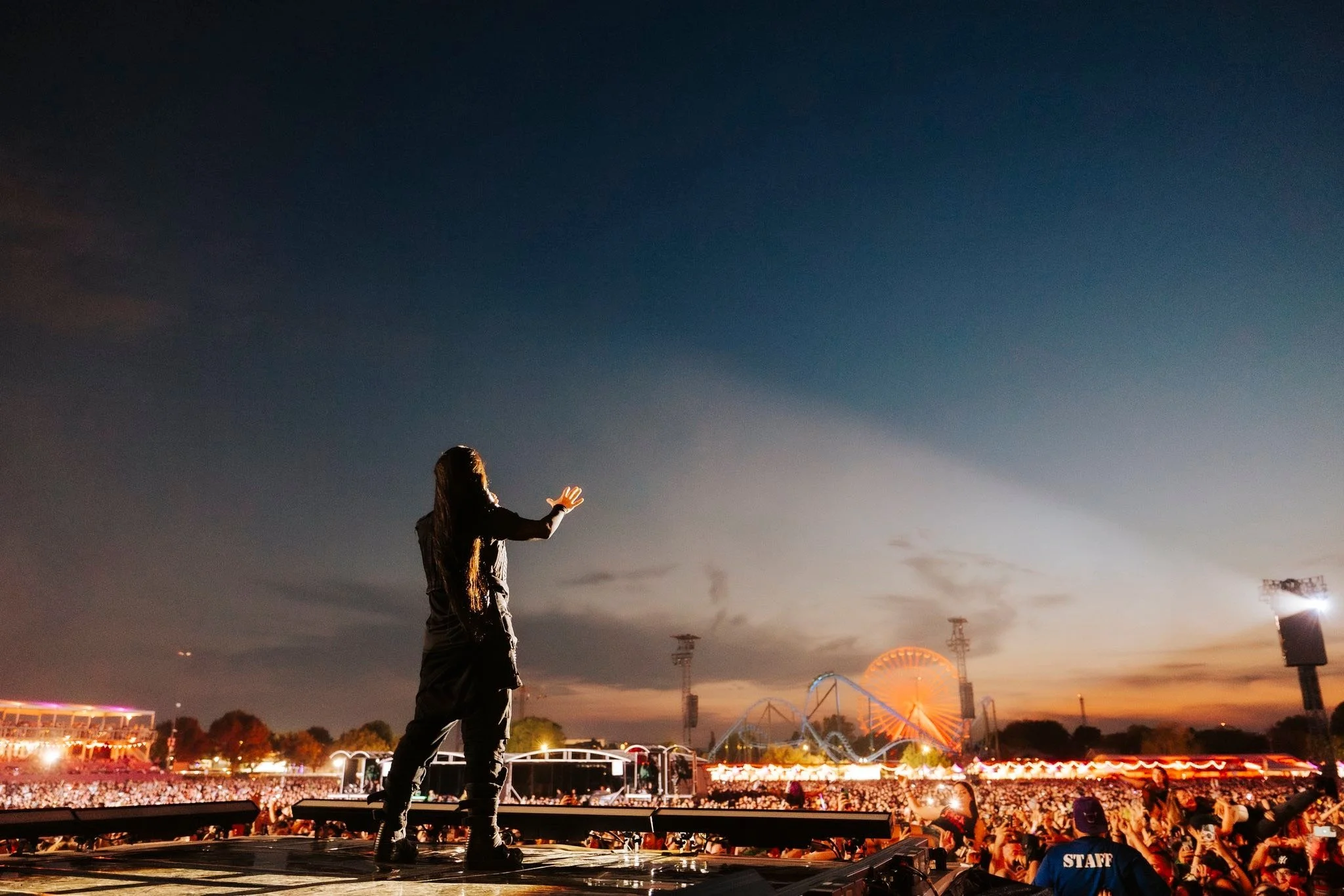 A woman on a stage at dusk with a large crowd in front, carnival rides and lights visible in the background.