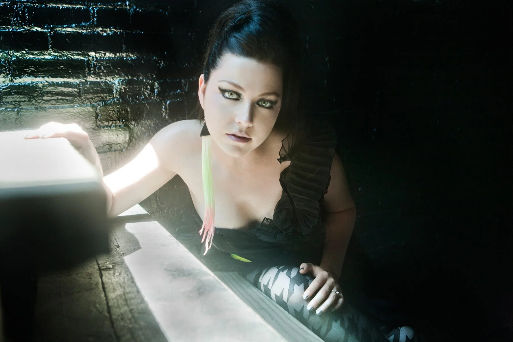 A woman with dark hair and striking makeup, sitting on a dark floor, with her upper body slightly above the ground, looking directly at the camera.