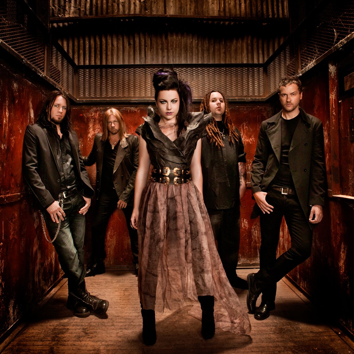 A gothic rock band in an industrial, rust-colored hallway. The lead woman wears a dark, ruffled top with a wide belt and flowing skirt, while the band members wear dark, edgy clothing with leather and metal accessories.
