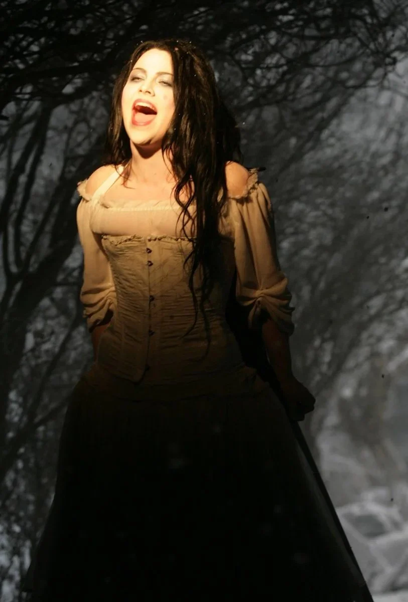 A woman with long dark hair, wearing a beige off-shoulder top, singing passionately with her mouth open, standing outdoors in a dark, wooded area.