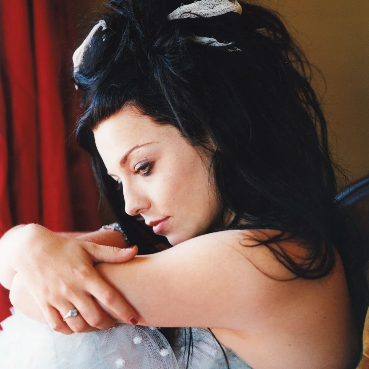 A woman with black, wavy hair and fair skin sitting indoors near a window with red curtains, resting her head on her arms, wearing a ring on her left hand, looking contemplative.