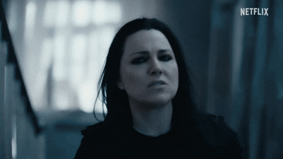 A woman with dark hair and serious expression looking forward, in a dimly lit room with curtains in the background.