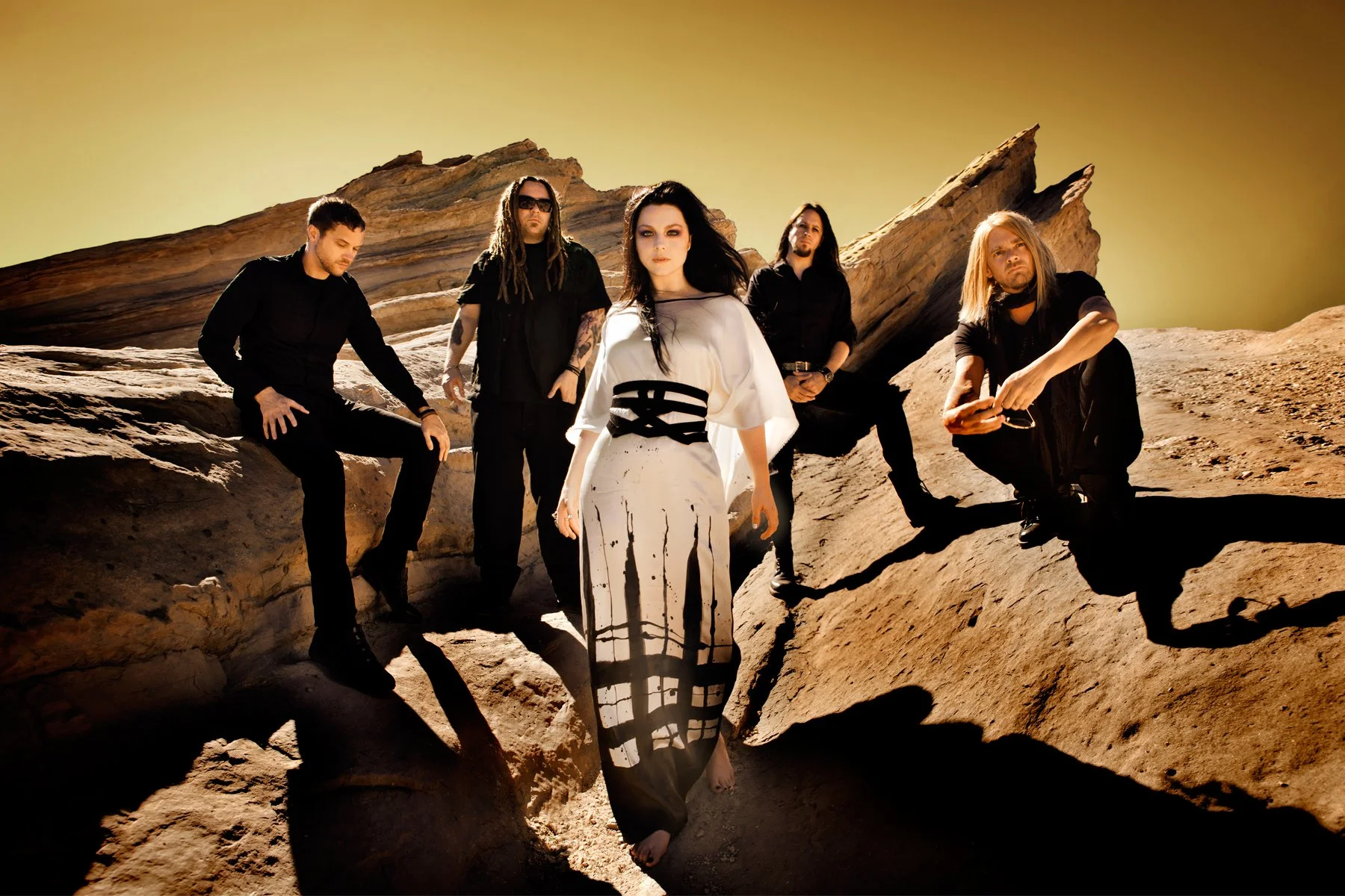 A band of five members dressed in black and white clothing poses on a rocky, desert-like landscape with large rock formations and a yellowish sky in the background.