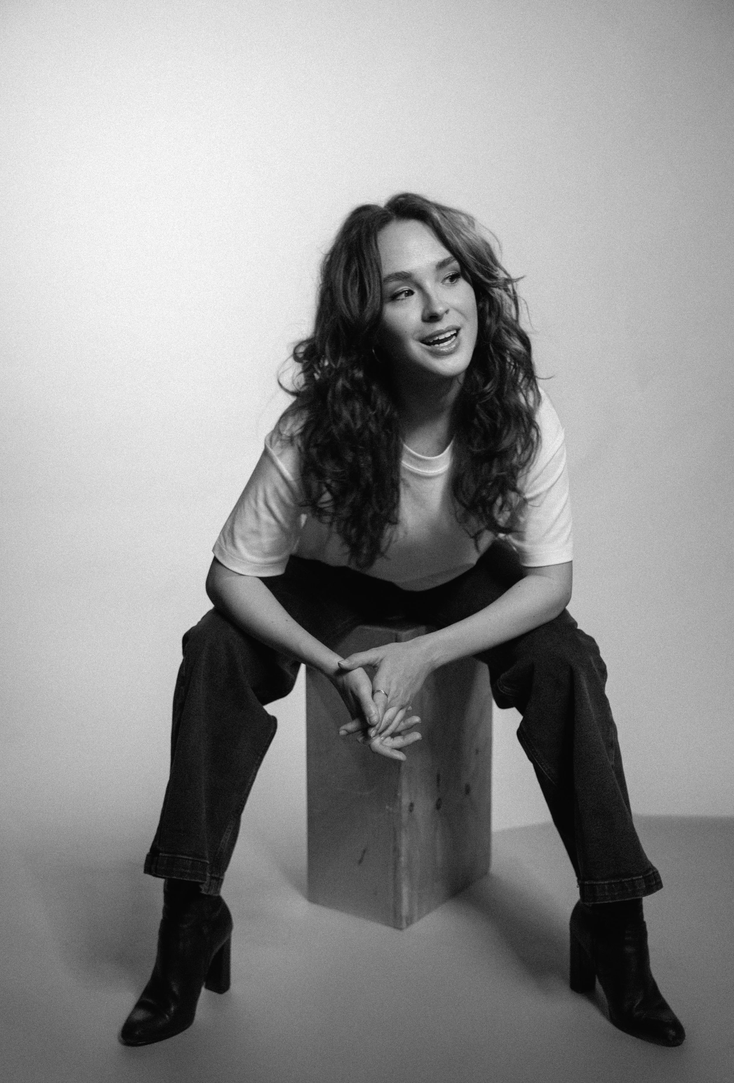 A woman with long, curly hair, wearing a white T-shirt and dark jeans, sitting on a wooden box with her legs apart and hands clasped, smiling and looking to her right, in a studio setting with a plain background.