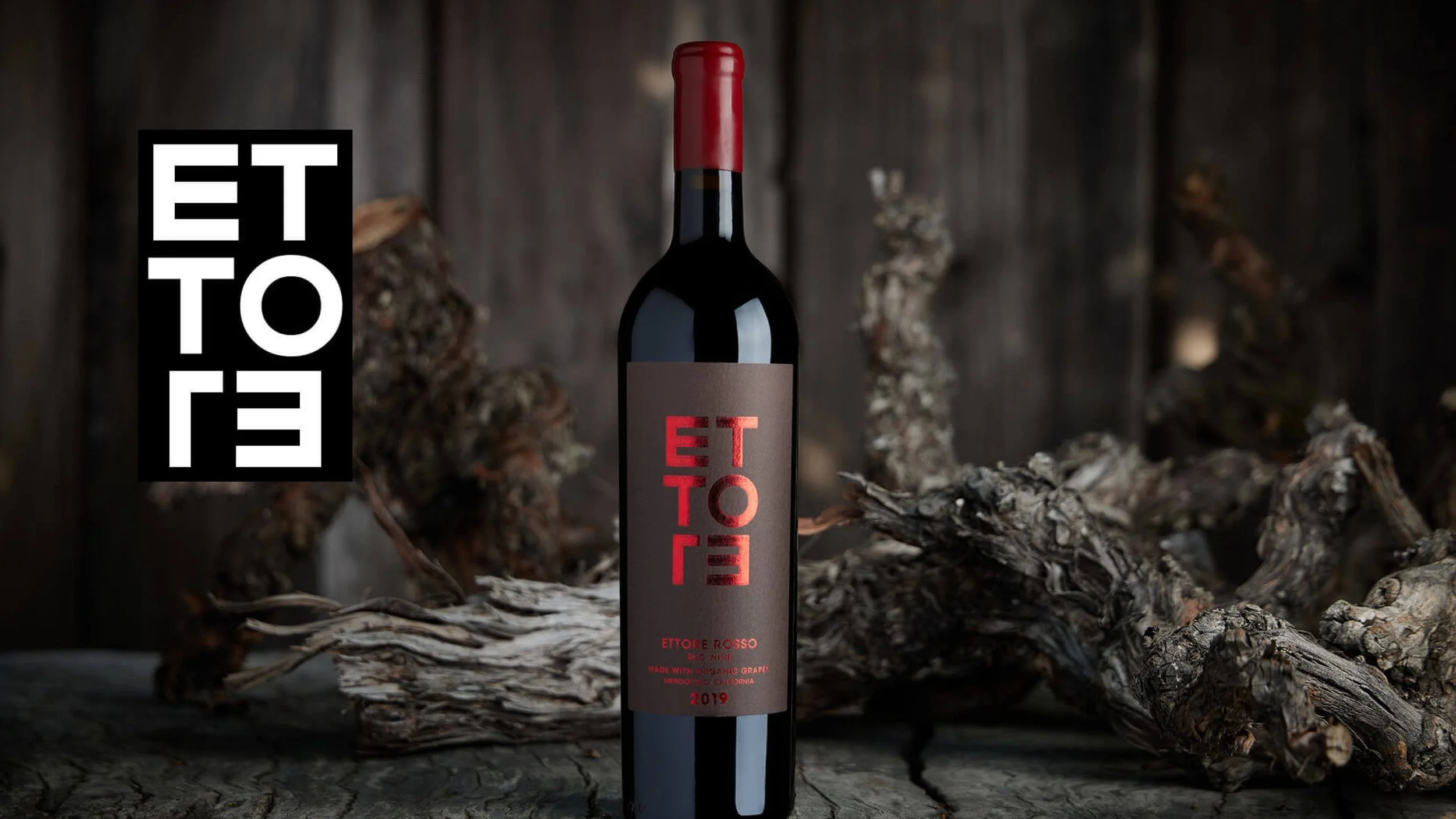 A bottle of red Ettore Rosso wine with a brown label that has red and black text, placed on a rustic wooden surface with twisted, dried grapevines in the background.