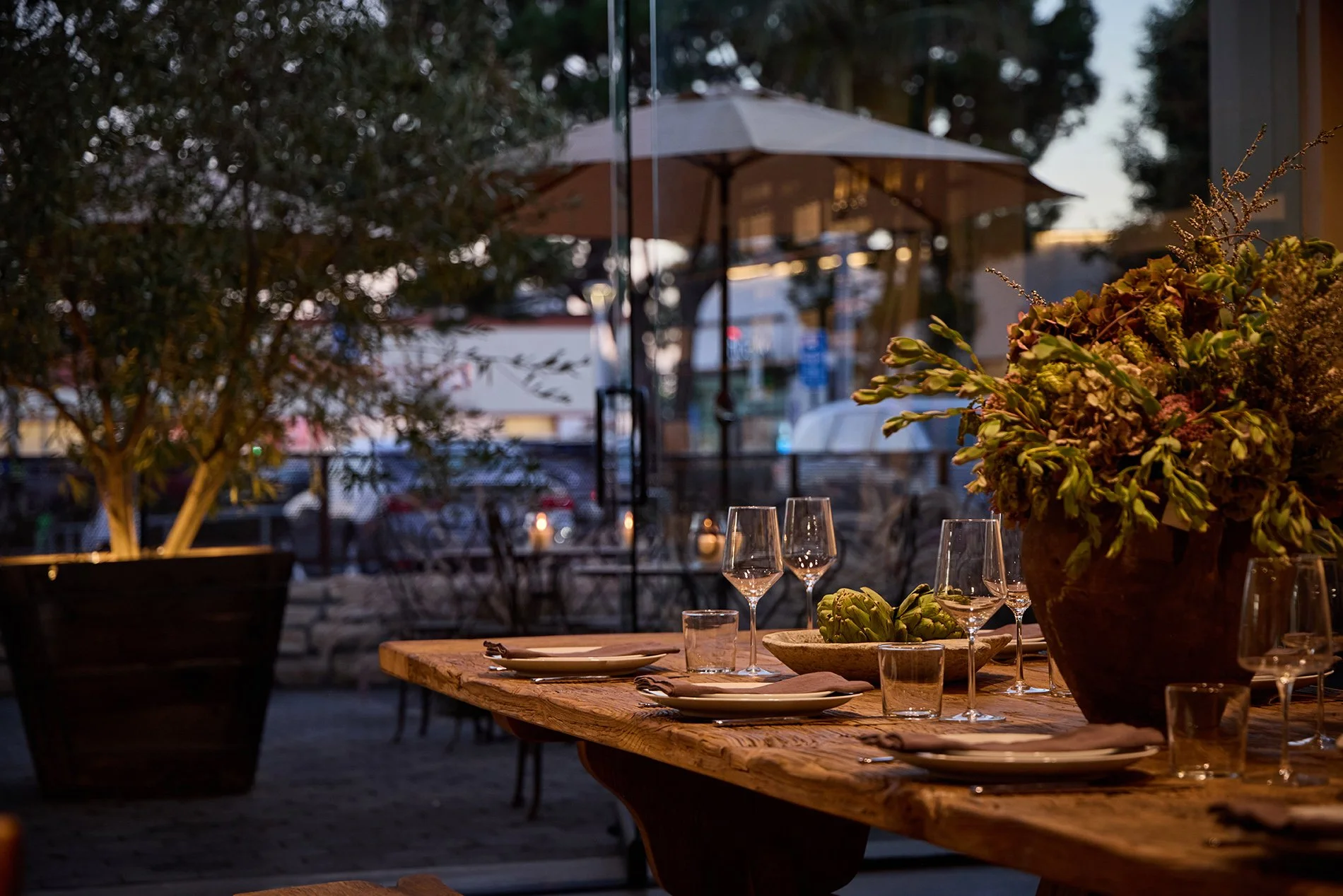 Outdoor dining table set for a meal with plates, glasses, and a large vase with flowers, under a patio umbrella at dusk.