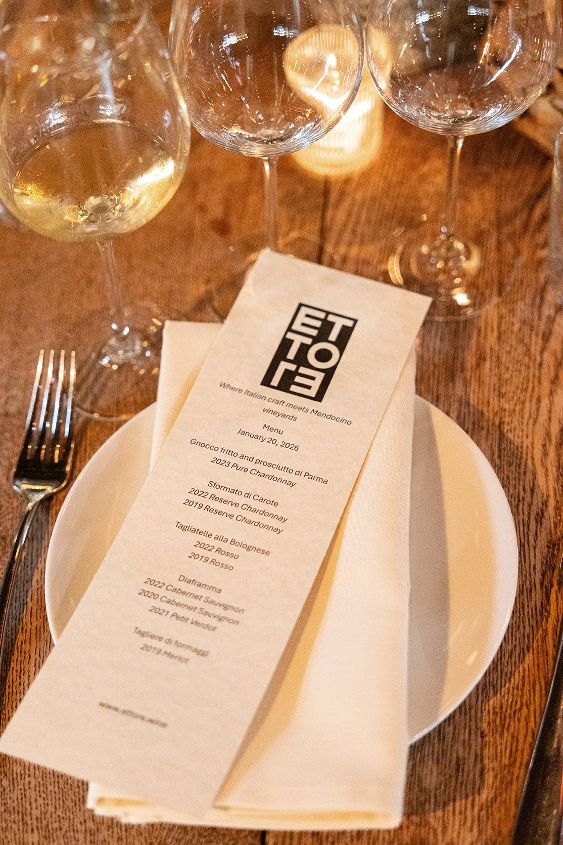 Wine glasses on a wooden table with a menu, plate, fork, and napkin.