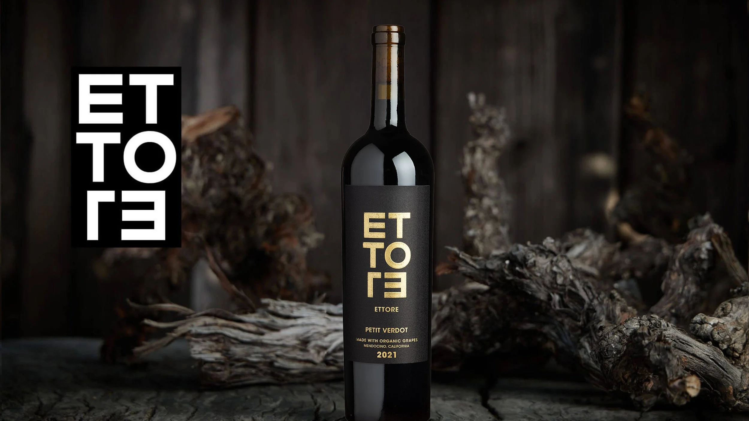 A bottle of ETTORE Petit Verdot wine from 2021, made with organic grapes from Mendocino, California, placed on a rustic wooden surface with dried vine and wood background.
