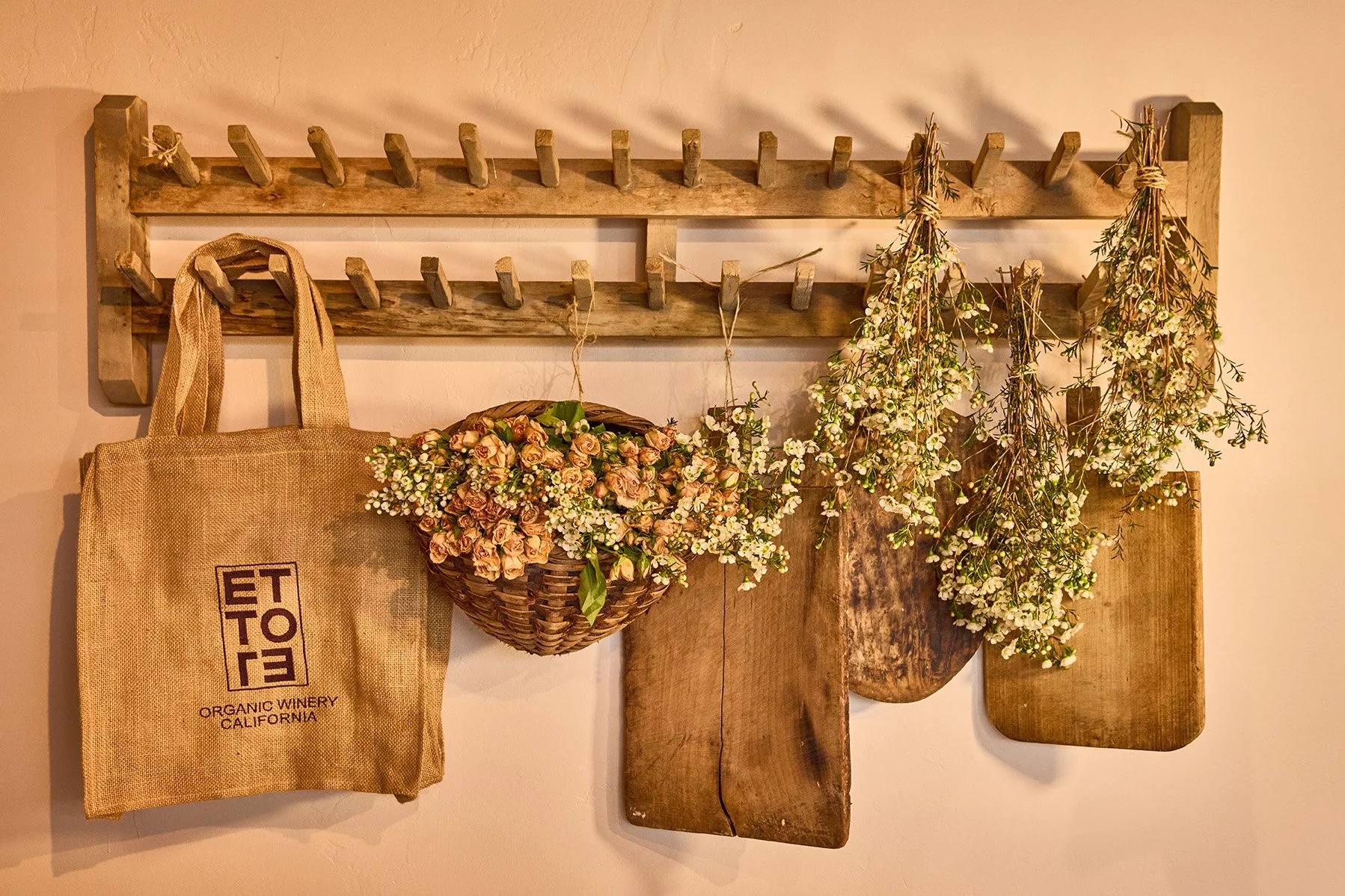 Wall hanging with wooden rack, a canvas bag with organic winery logo, a basket of pink and white flowers, and hanging sprigs of small white flowers and greenery.