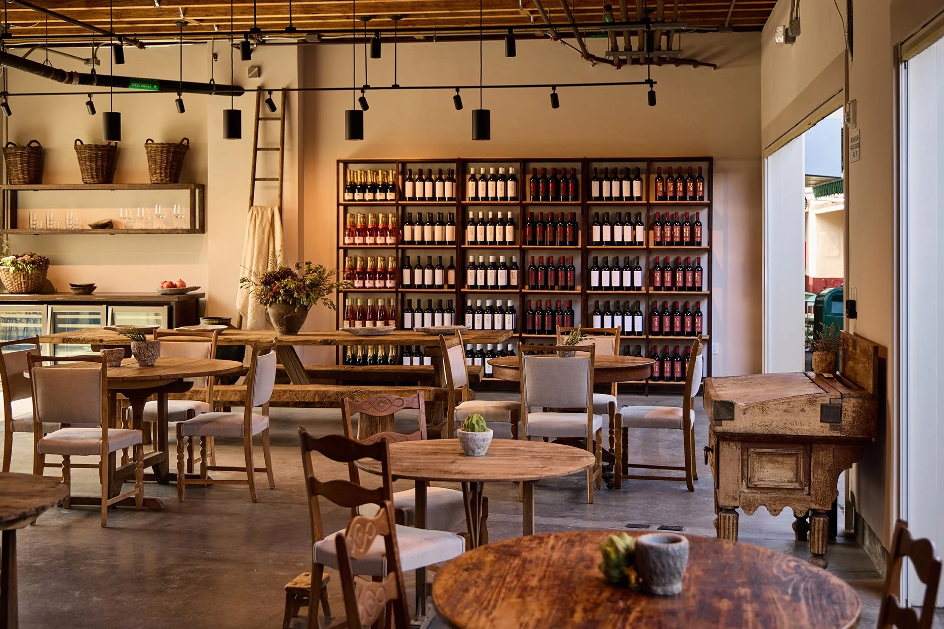 Interior of a cozy cafe or restaurant with wooden tables and chairs, a wall with wine bottles on shelves, and decorative plants on the tables.
