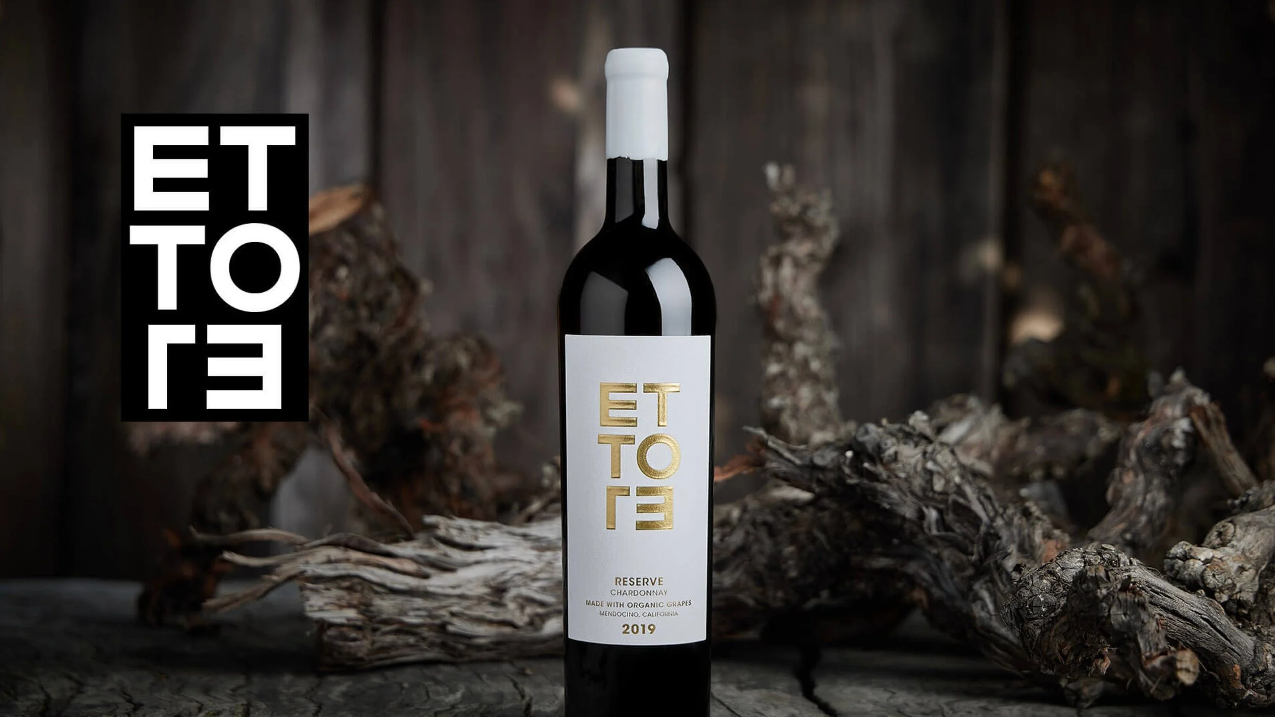 A bottle of ETTOE Reserve Chardonnay 2019 wine positioned on a textured surface with twisted, weathered wood pieces around it, against a dark wooden background.