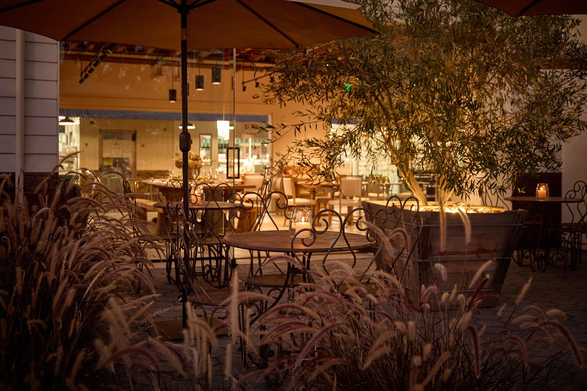 Outdoor patio at Ettore Osteria at night with lit candles, chairs, tables, large potted plant, and decorative grasses.
