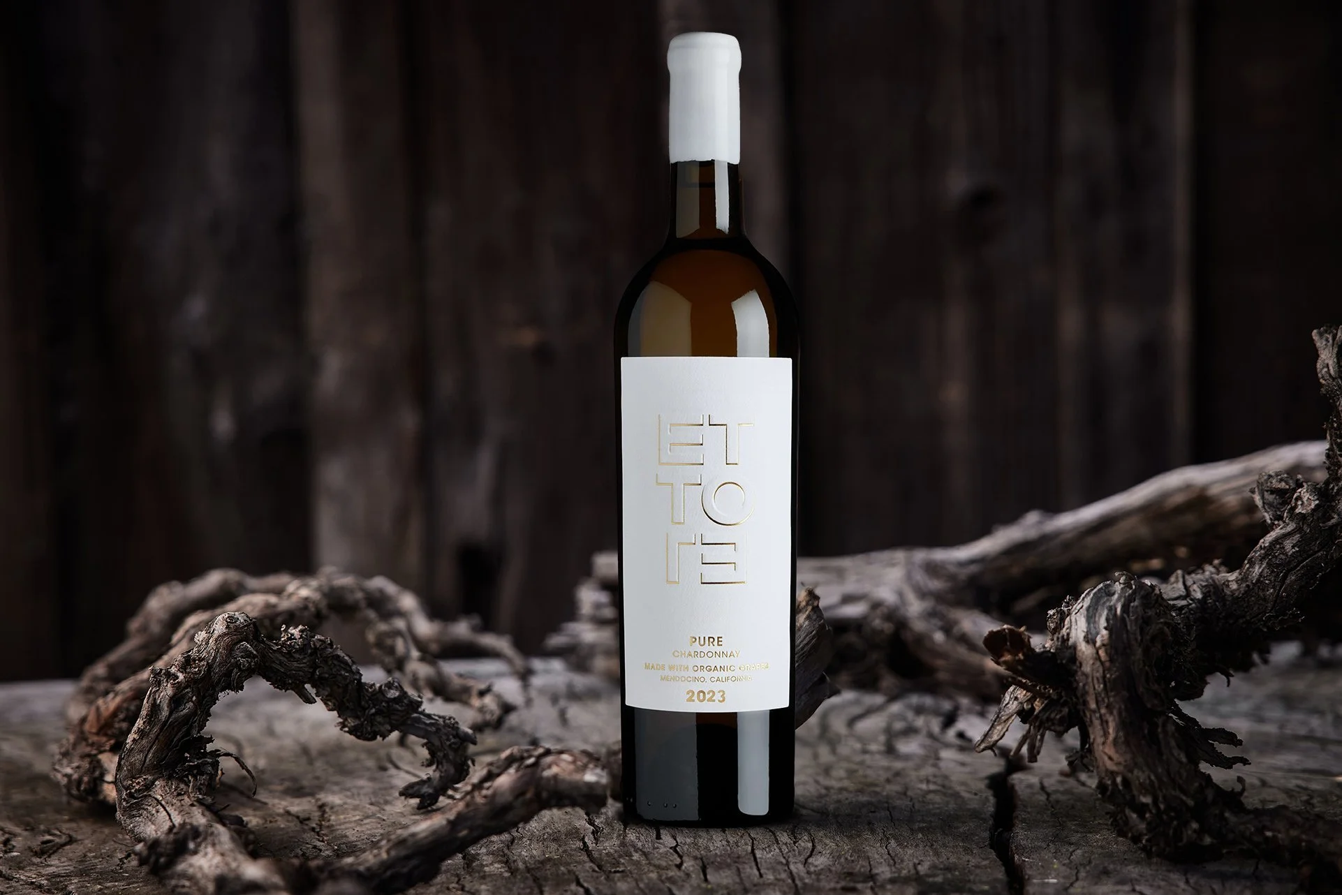 A bottle of white wine labeled '2023 Ettore Pure Chardonnay' with organic grapes, vintage 2023, standing on a rustic wooden surface with twisted branches in the foreground and a dark wooden background.