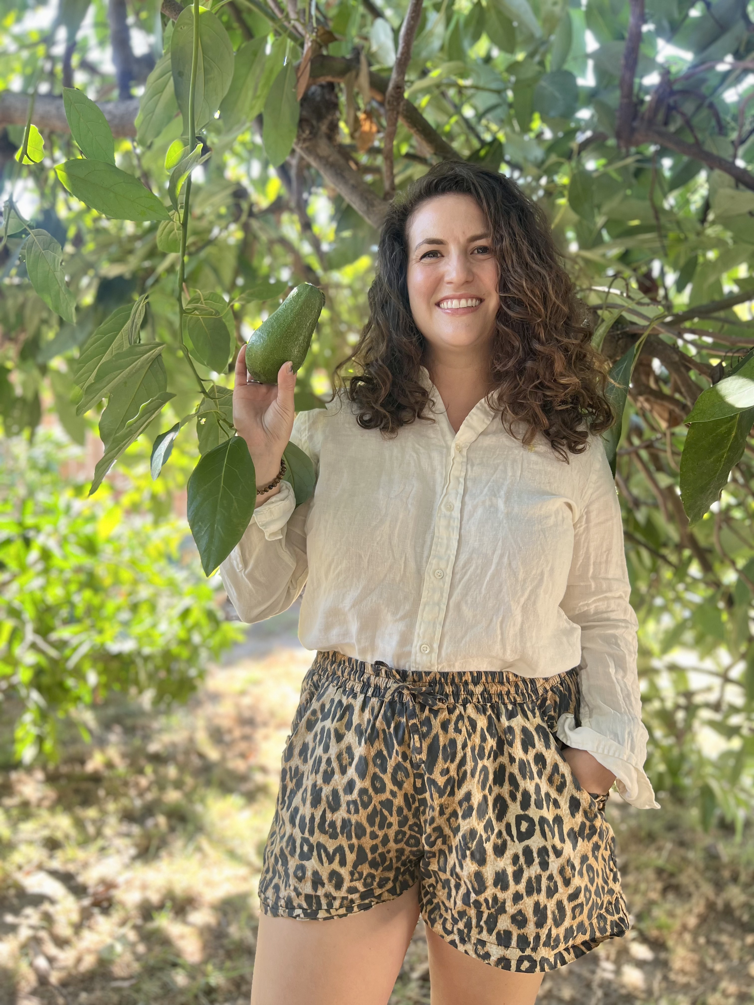 Smiling woman with curly hair holding an avocado in a lush green outdoor setting.