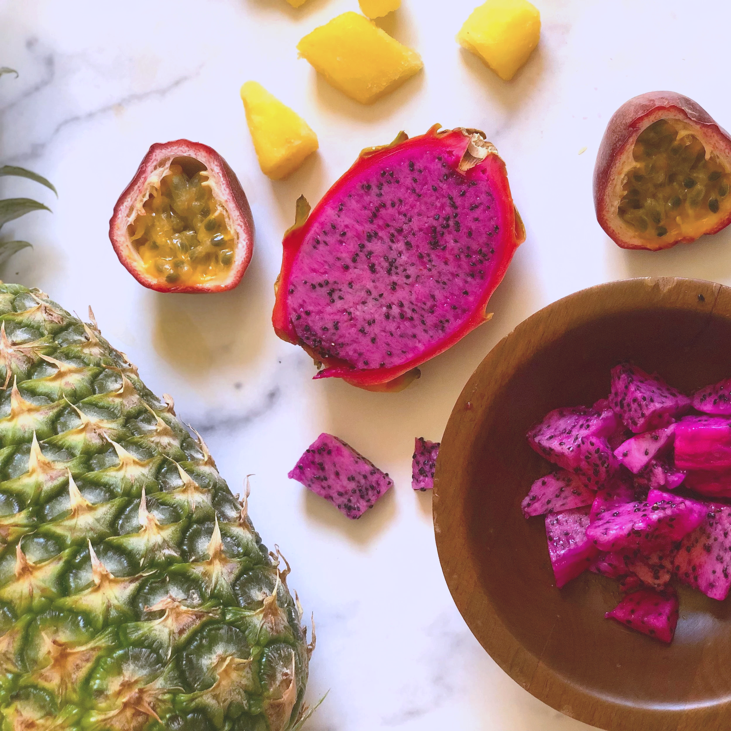 Fresh pineapple, passion fruit, dragon fruit, pineapple chunks, and a bowl of diced dragon fruit on a white surface.