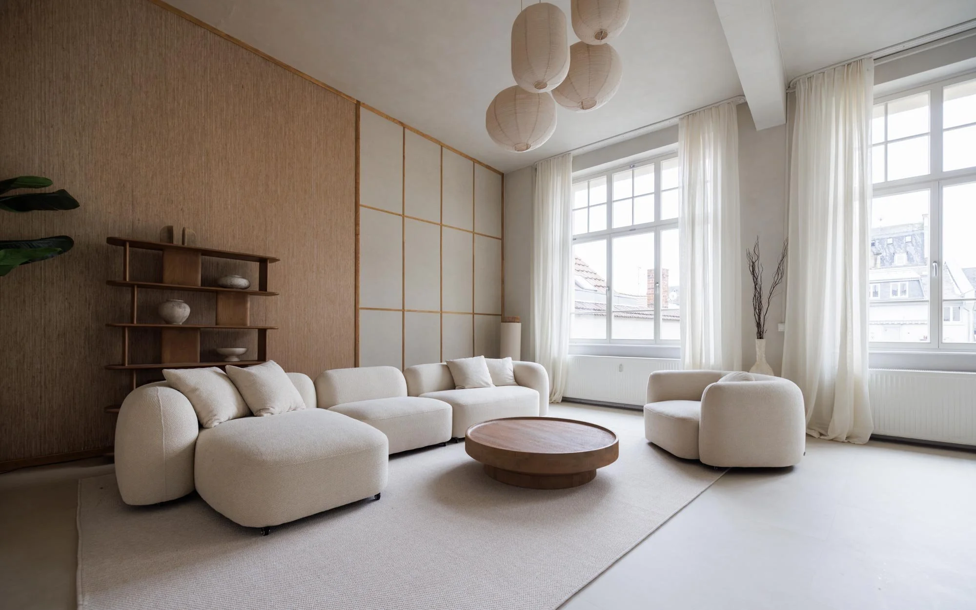 Helles Wohnzimmer mit großen Fenstern, weißen Vorhängen, einer weißen Couch, einem weißen Sessel, einem runden Holztisch, einer Wand mit Holzregalen und Dekorationen, und einer stilvollen Deckenleuchte.