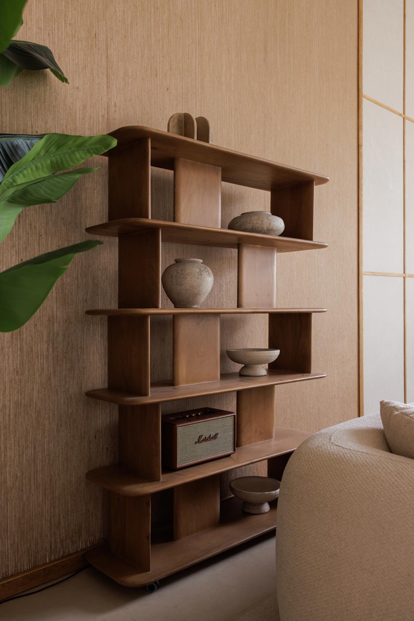 Ein Holzregal mit mehreren Fächern, darauf befinden sich keramische Dekorationsgefäße und ein kleiner Marshall-Lautsprecher. Das Regal steht an einer Wand mit Tapete, neben einer Couch und einer großen Pflanze.