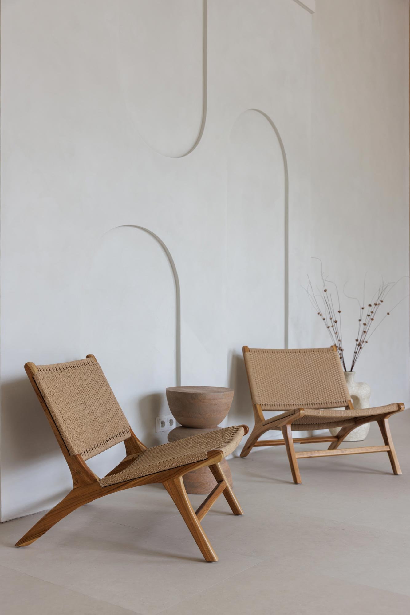 Zwei Holzsessel mit geflochtenen Sitzen vor einer weißen Wand mit Zierleisten, eine Vase mit Zweigen im Hintergrund, ein runder Holztisch zwischen den Sesseln.