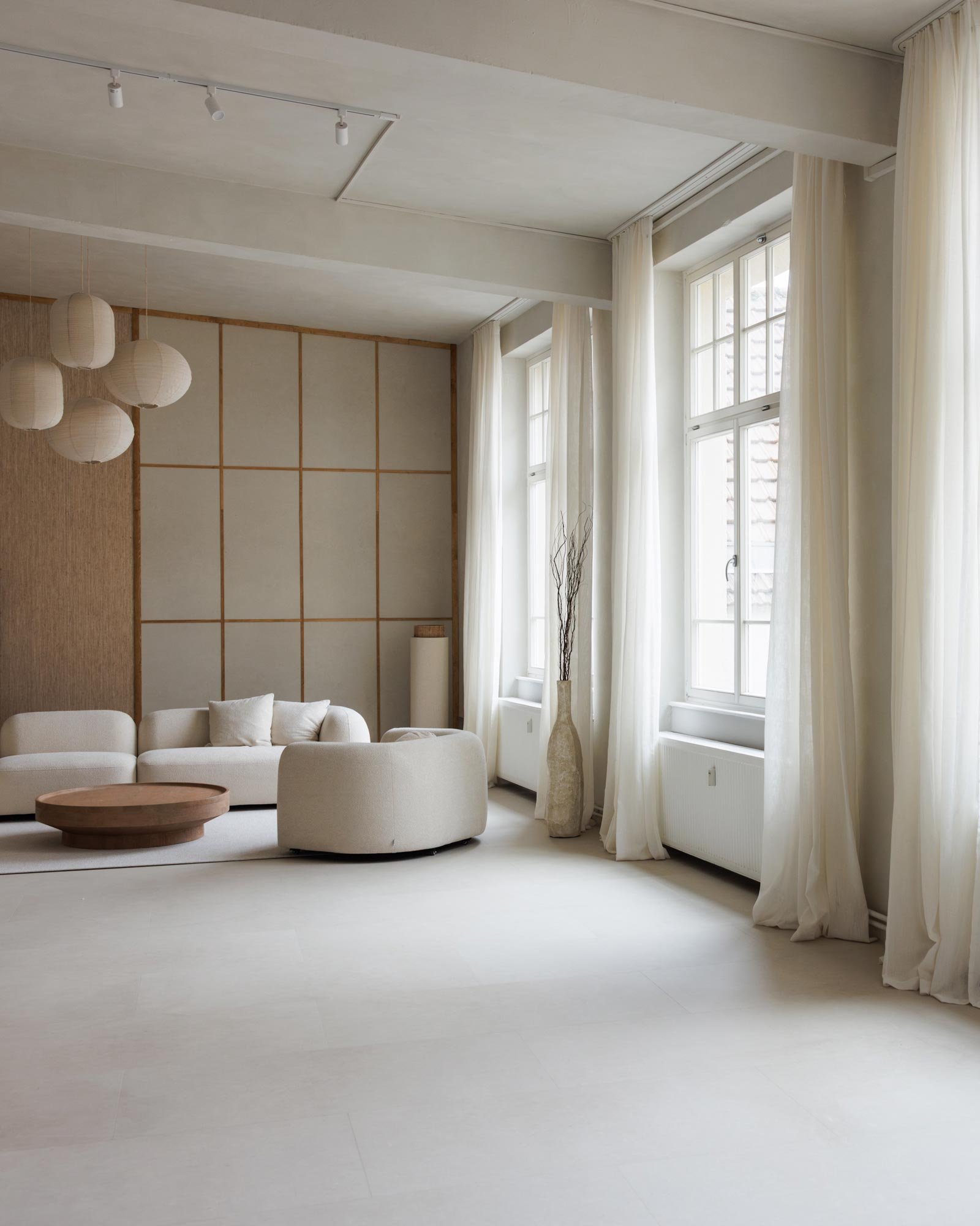 Ein helles, minimalistisch eingerichtetes Wohnzimmer mit großen Fenstern, weißen Vorhängen, weißem Sofa, einem runden Holz Couchtisch und einer dekorativen Vase mit Zweigen.