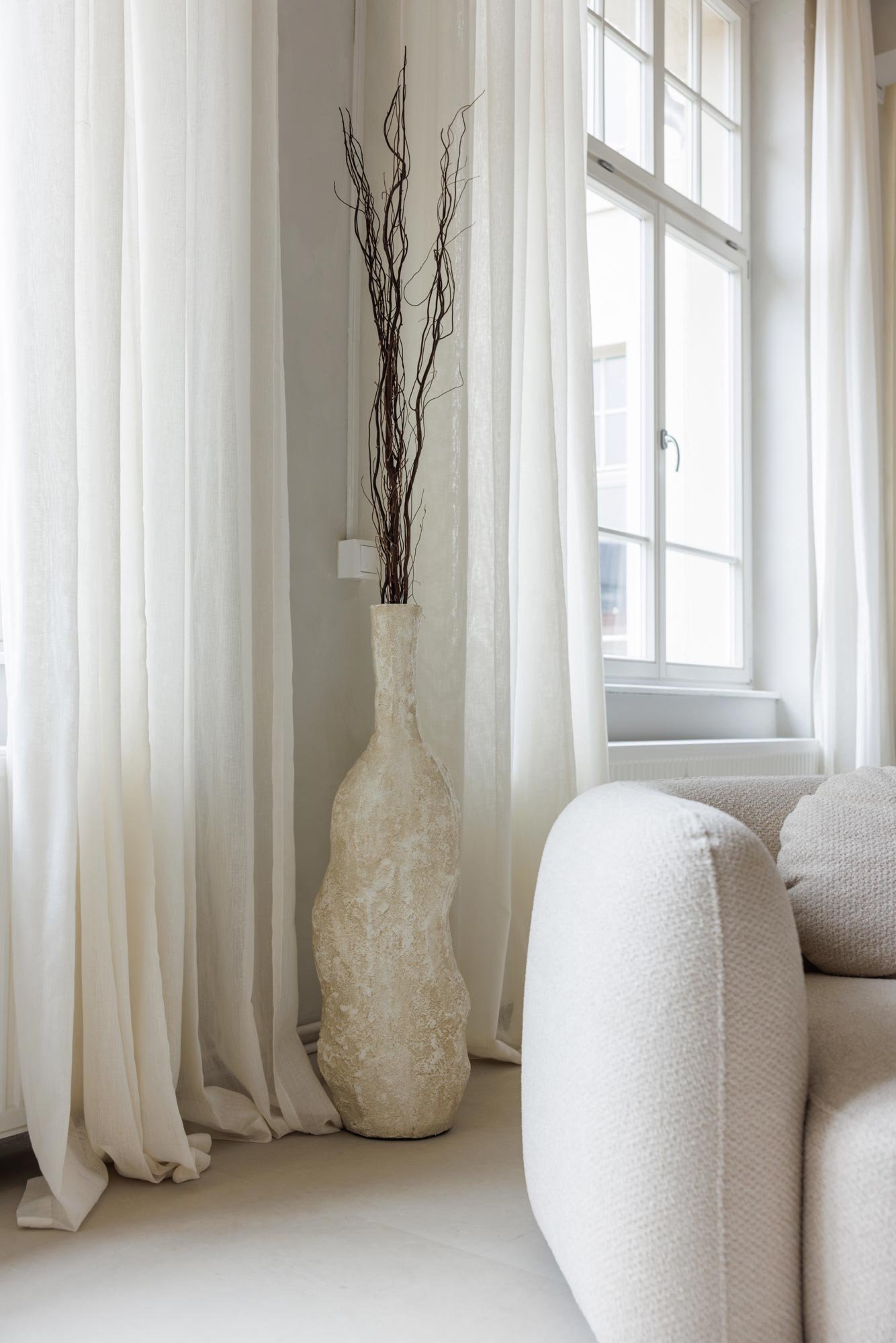 Eine weiße, grob strukturierte Vase mit langen, dünnen Zweigen steht neben einem Fenster in einem hellen Wohnzimmer.