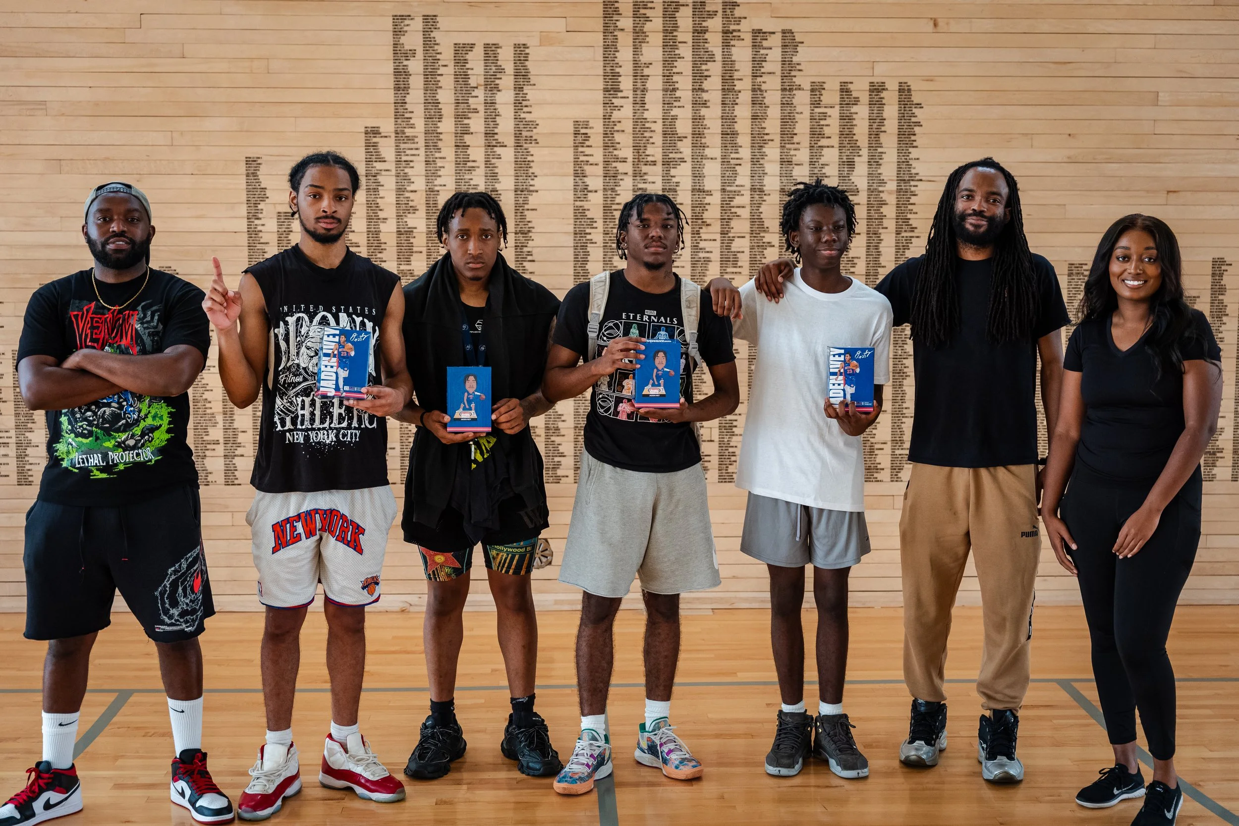 Group of seven young adults standing on a basketball court, holding awards or trophies, in front of a wall with a large memorial or list of names.