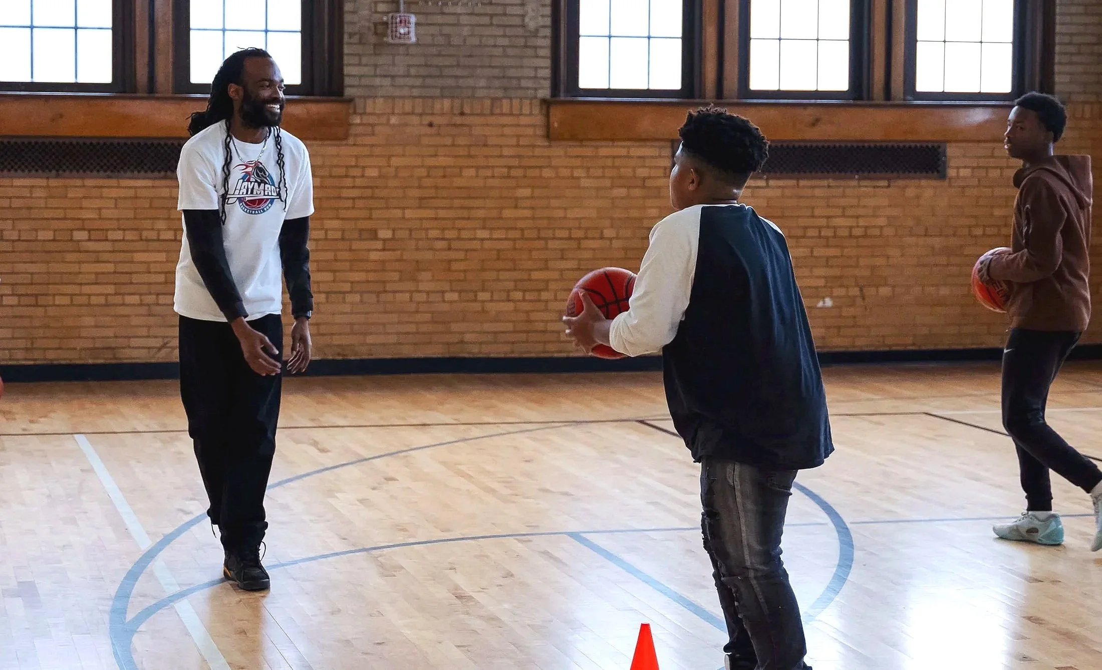 A basketball coach and two young players practicing in an indoor gym with brick walls. The player in the foreground is holding a basketball, and the coach is smiling.