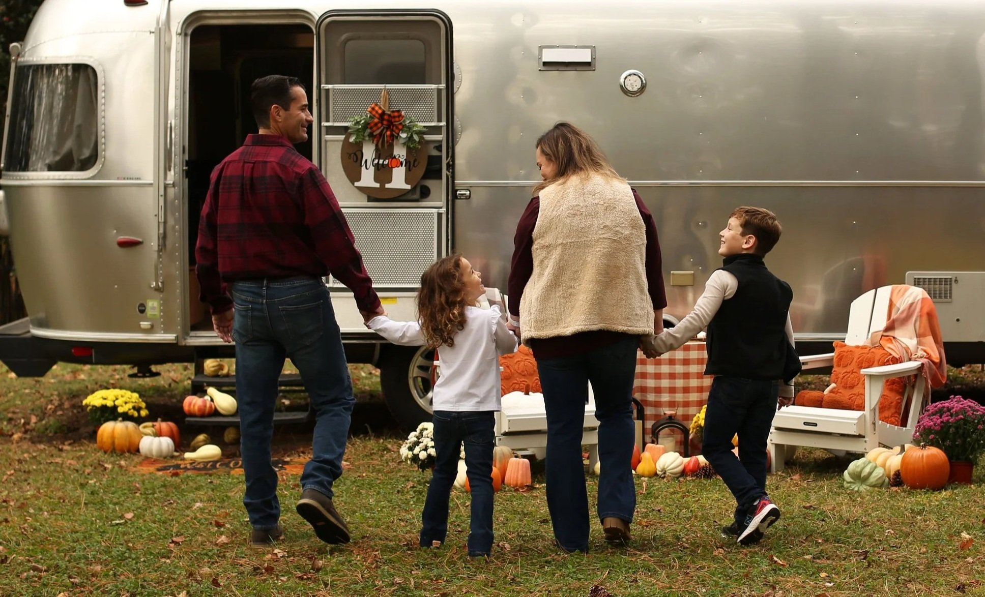 A family of four holding hands, walking in front of a silver camper trailer decorated with fall pumpkins and flowers. They are outdoors on a grassy area.