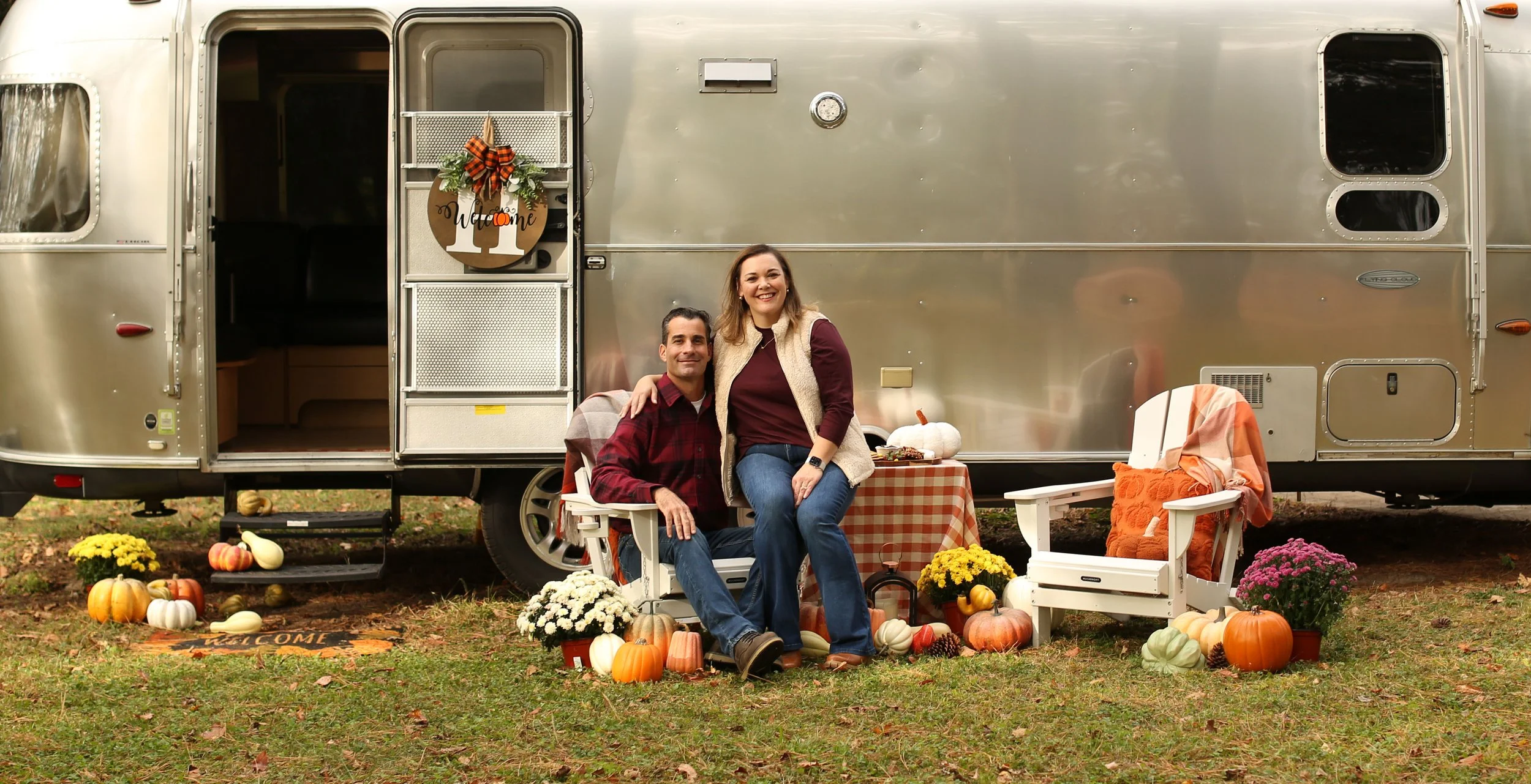 A smiling couple sitting outside an Airstream trailer decorated for fall with pumpkins, gourds, and flowers, surrounded by autumnal decorations and seating area.