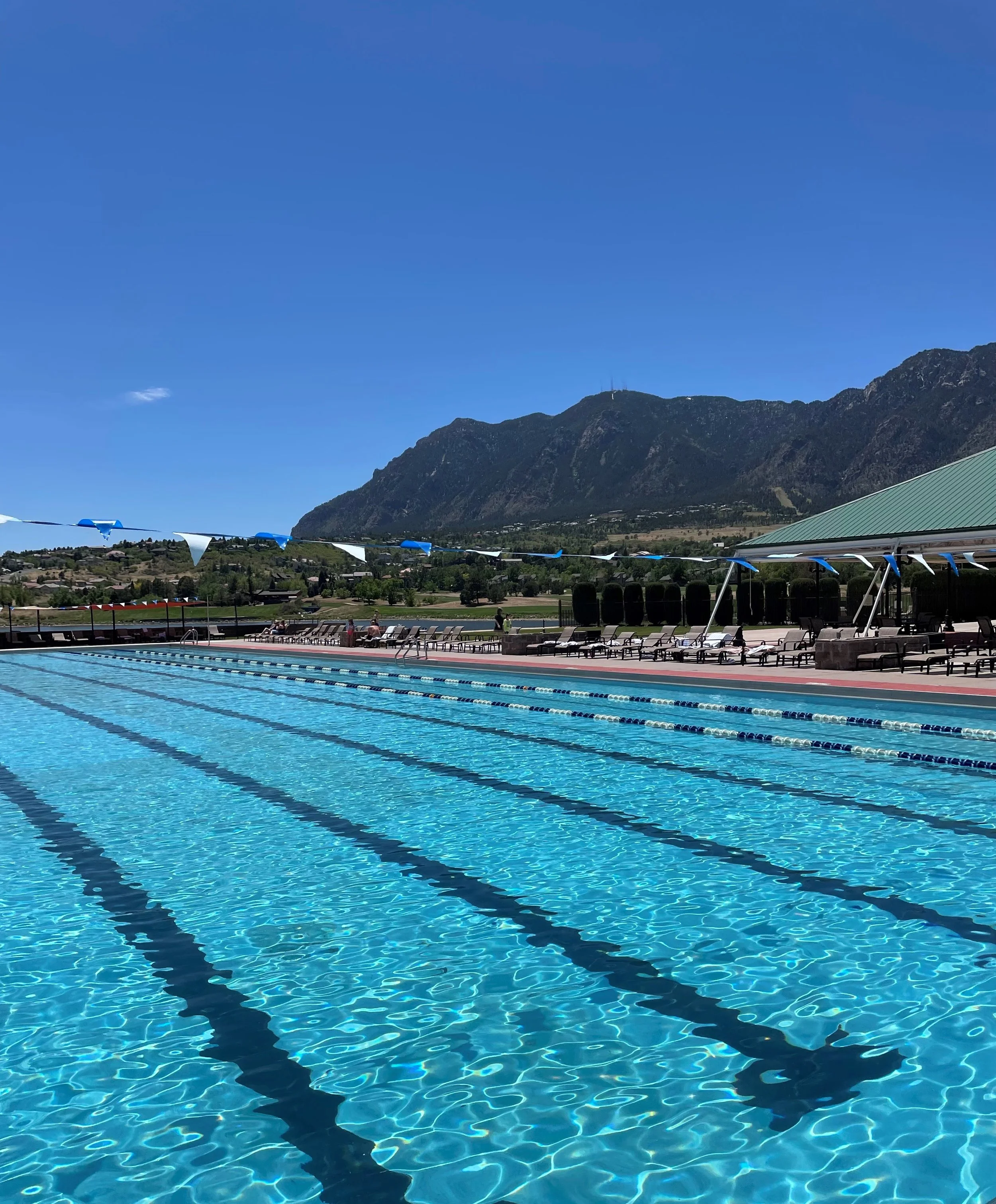 Outdoor swimming pool with lane dividers, surrounded by lounge chairs and a shaded area, with mountains and a clear blue sky in the background.