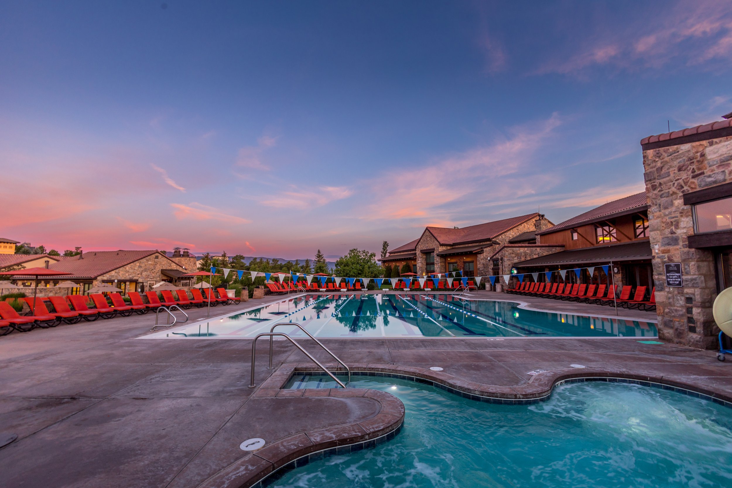 An outdoor swimming pool and hot tub area at sunset with empty red lounge chairs lining the pool and a stone building in the background.