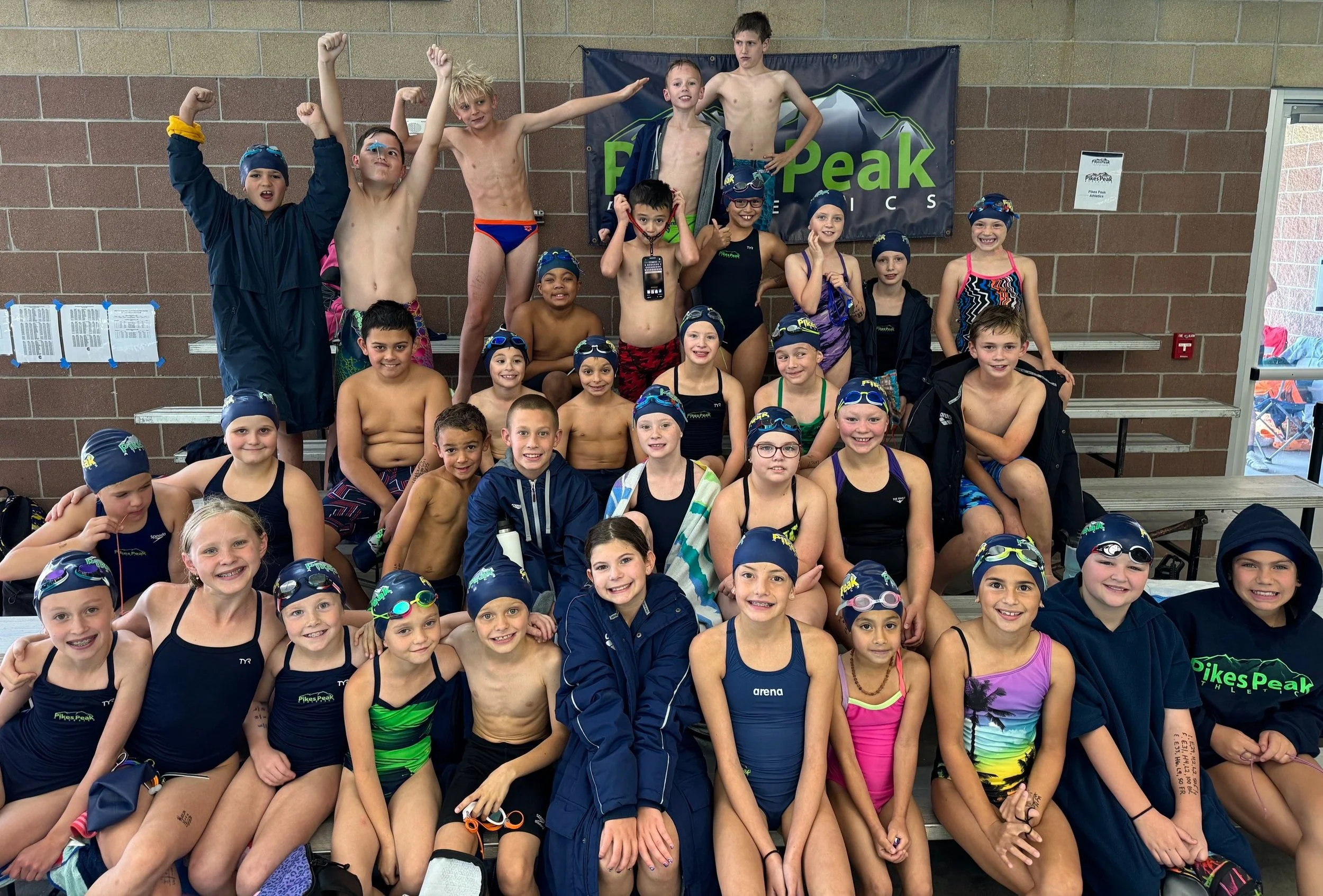 Group of young swimmers and their coach at an indoor swimming pool, posing for a group photo. Many are wearing swim caps, goggles, and swimsuits, smiling and showing excitement.