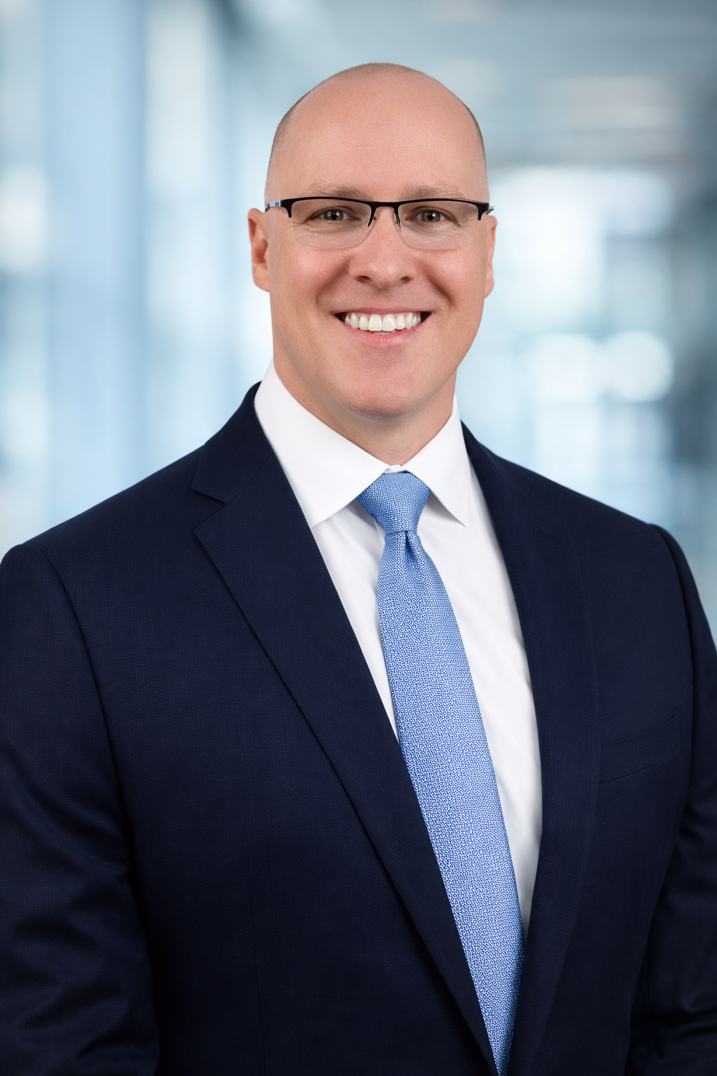 Professional headshot of a smiling man in glasses, navy suit, white shirt, and light blue tie, with a blurred office background.