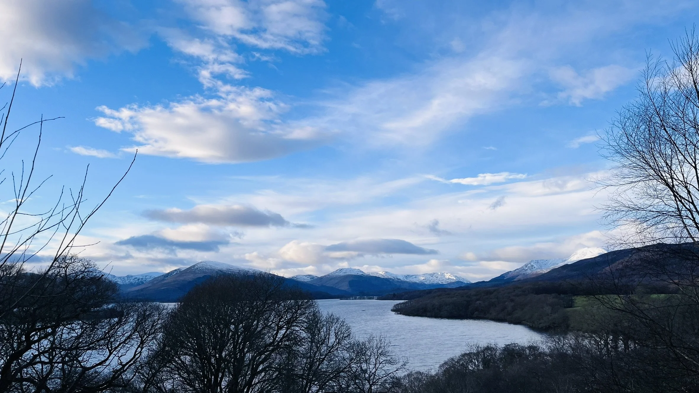 A landscape featuring a river winding through mountains with snow-capped peaks in the background, under a partly cloudy sky, with leafless trees in the foreground.