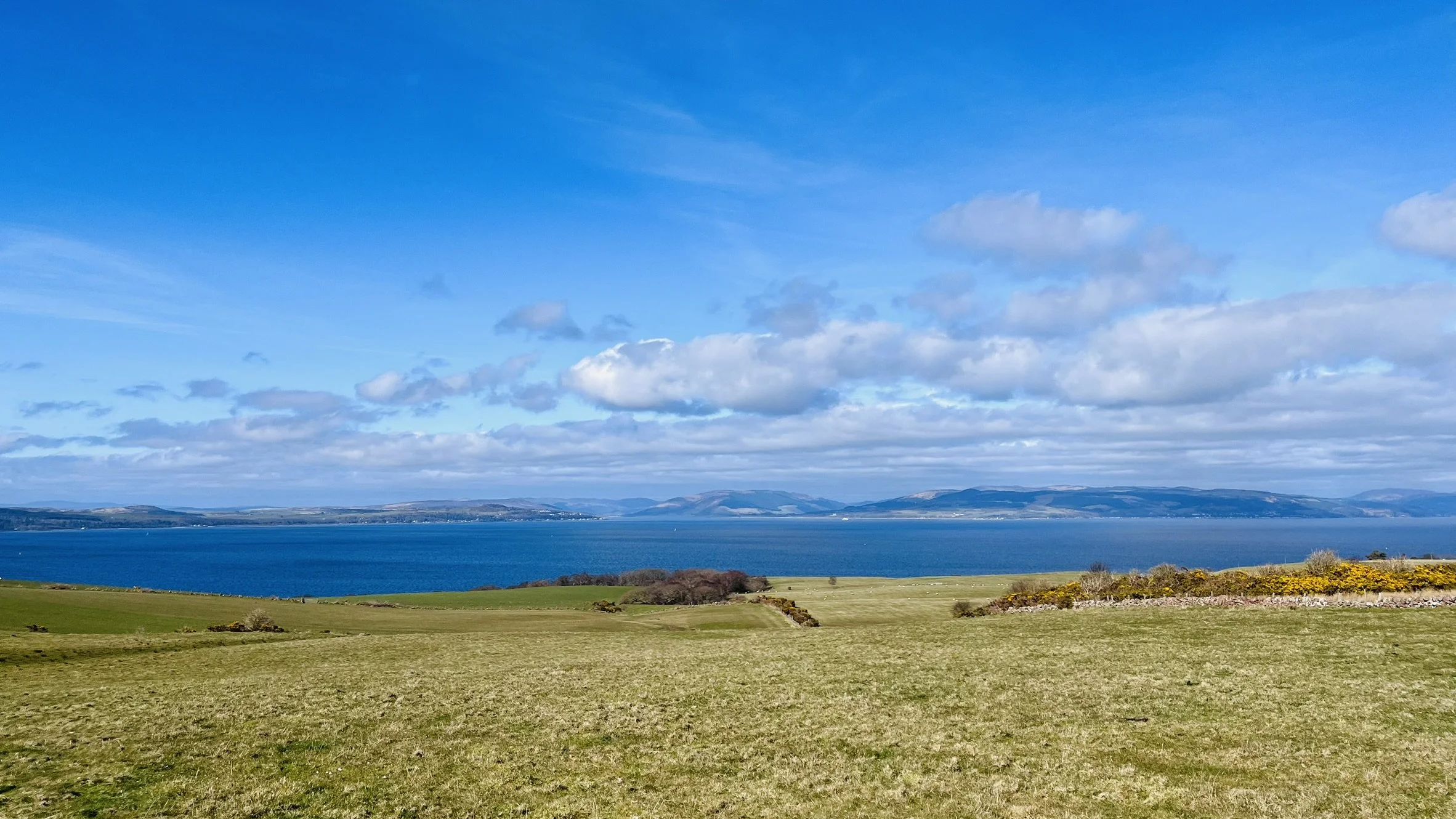 Open grassy field leading to a large blue lake with hills and mountains in the distance under a partly cloudy sky.