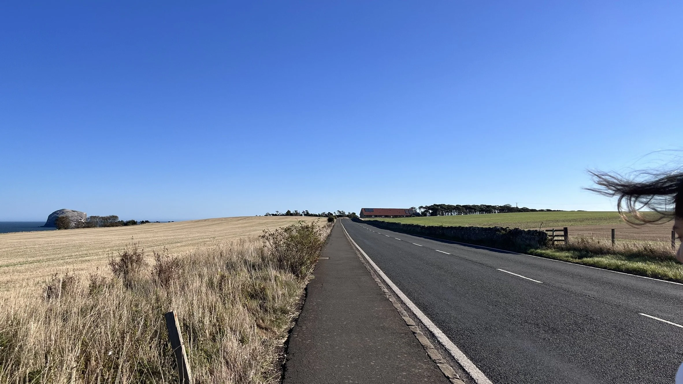 A rural road running through open fields with a rocky hill in the distance under a clear blue sky, with a person on the right side of the image.