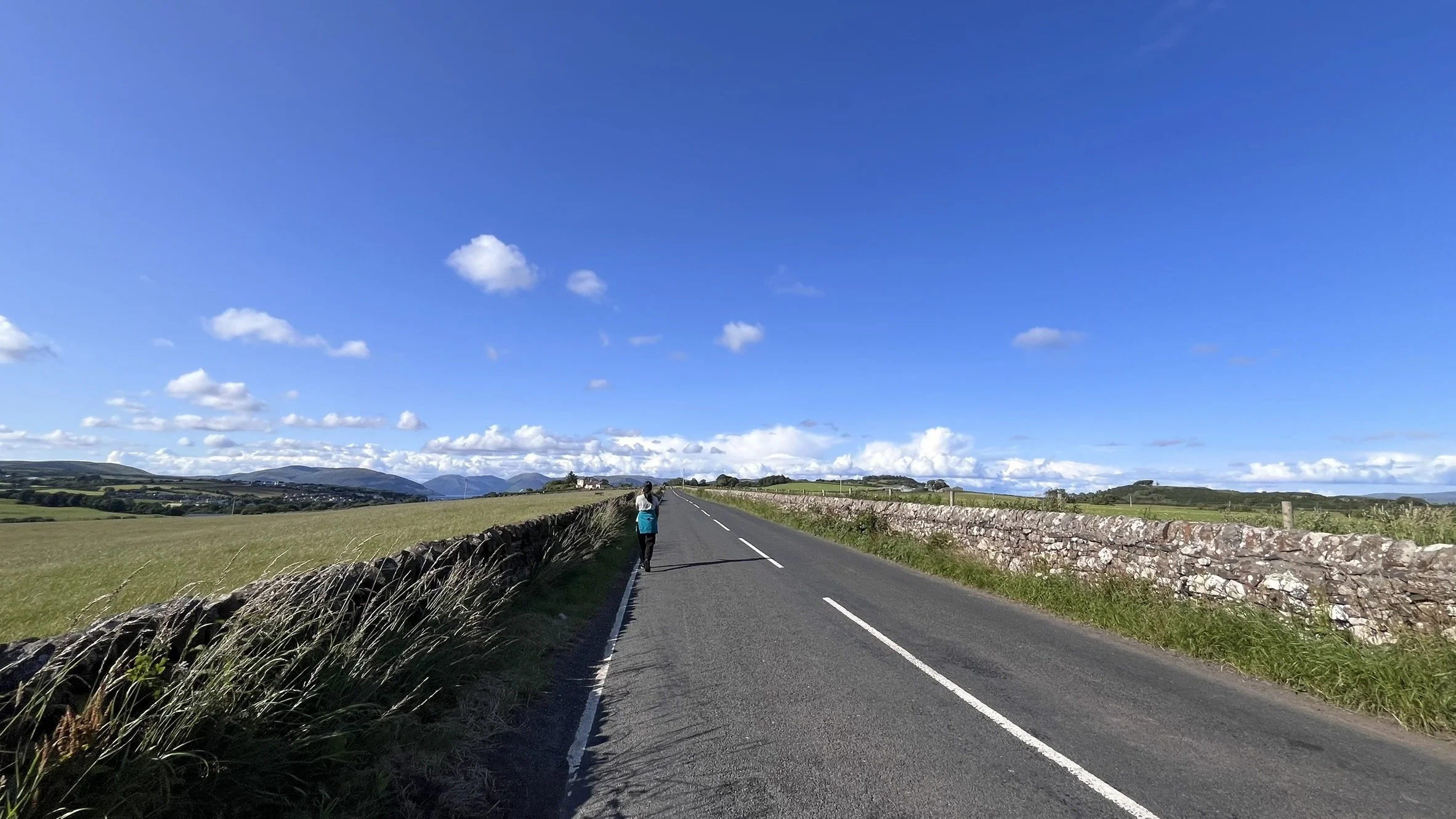 A person walking on a rural road with stone walls on both sides, under a blue sky with scattered clouds, landscape of green fields and distant hills.