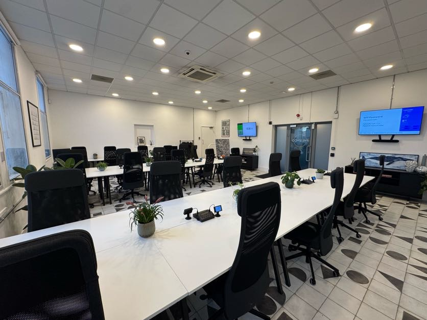 Conference room with large white table, multiple black chairs, potted plants, and large monitors on the walls.
