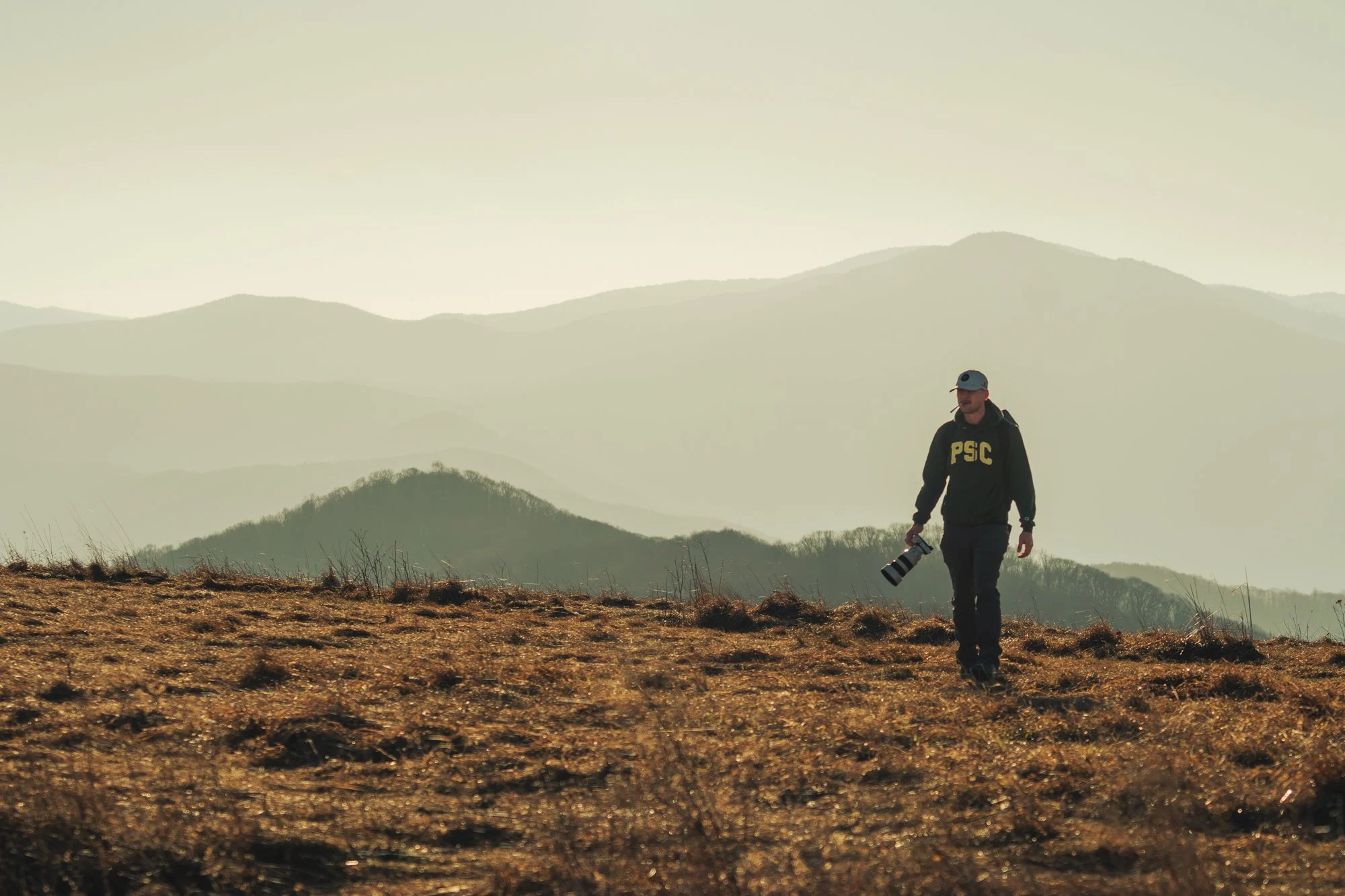 A person walking in a field holding a camera, with mountains in the background and a hazy sky.